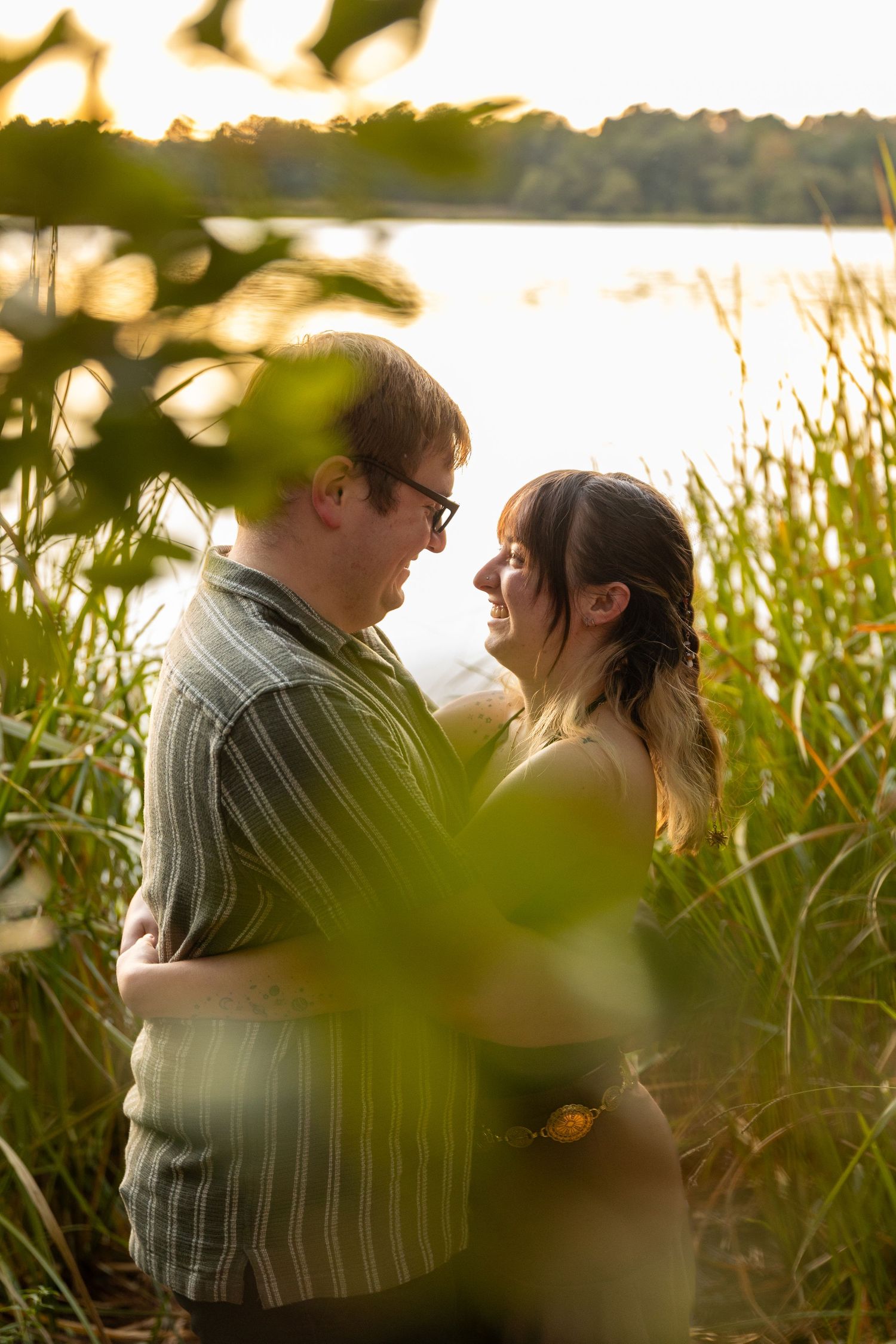 Intimate sunset photoshoot of a couple embracing among tall grass near a lake.