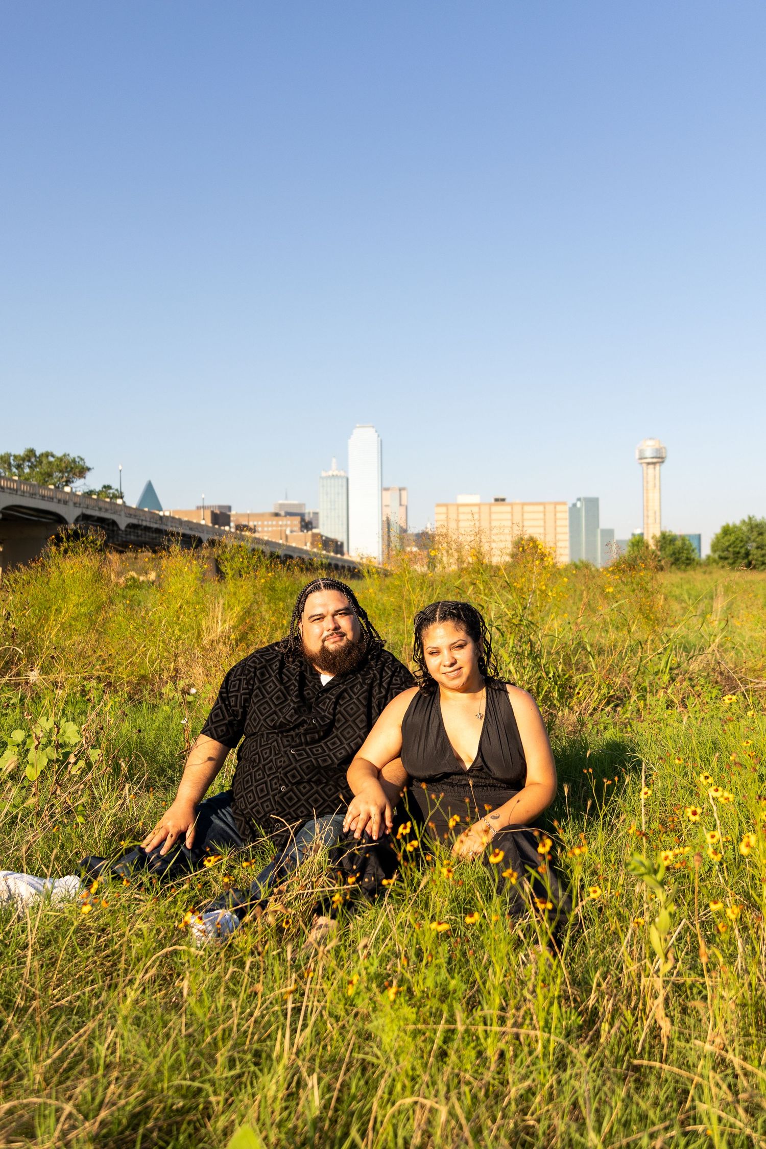 Couple lounging in tall grass with city skyline visible in background during sunset.