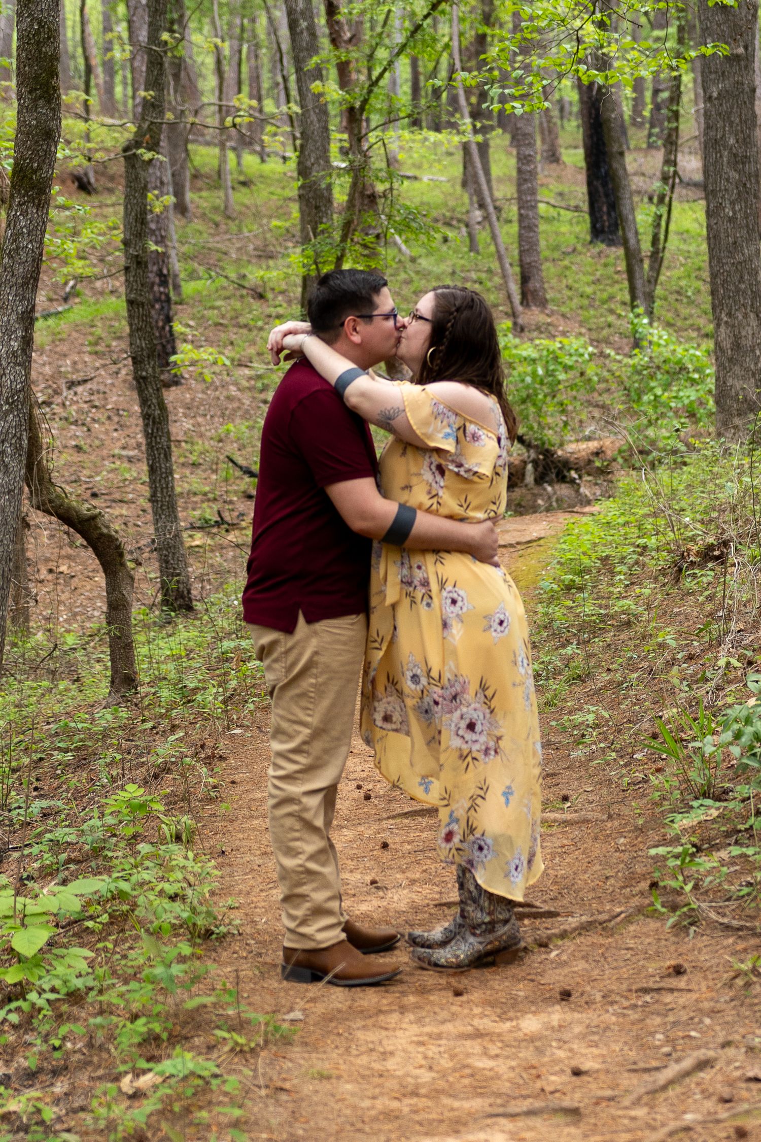 A couple embraces on a wooded trail surrounded by trees and natural scenery.