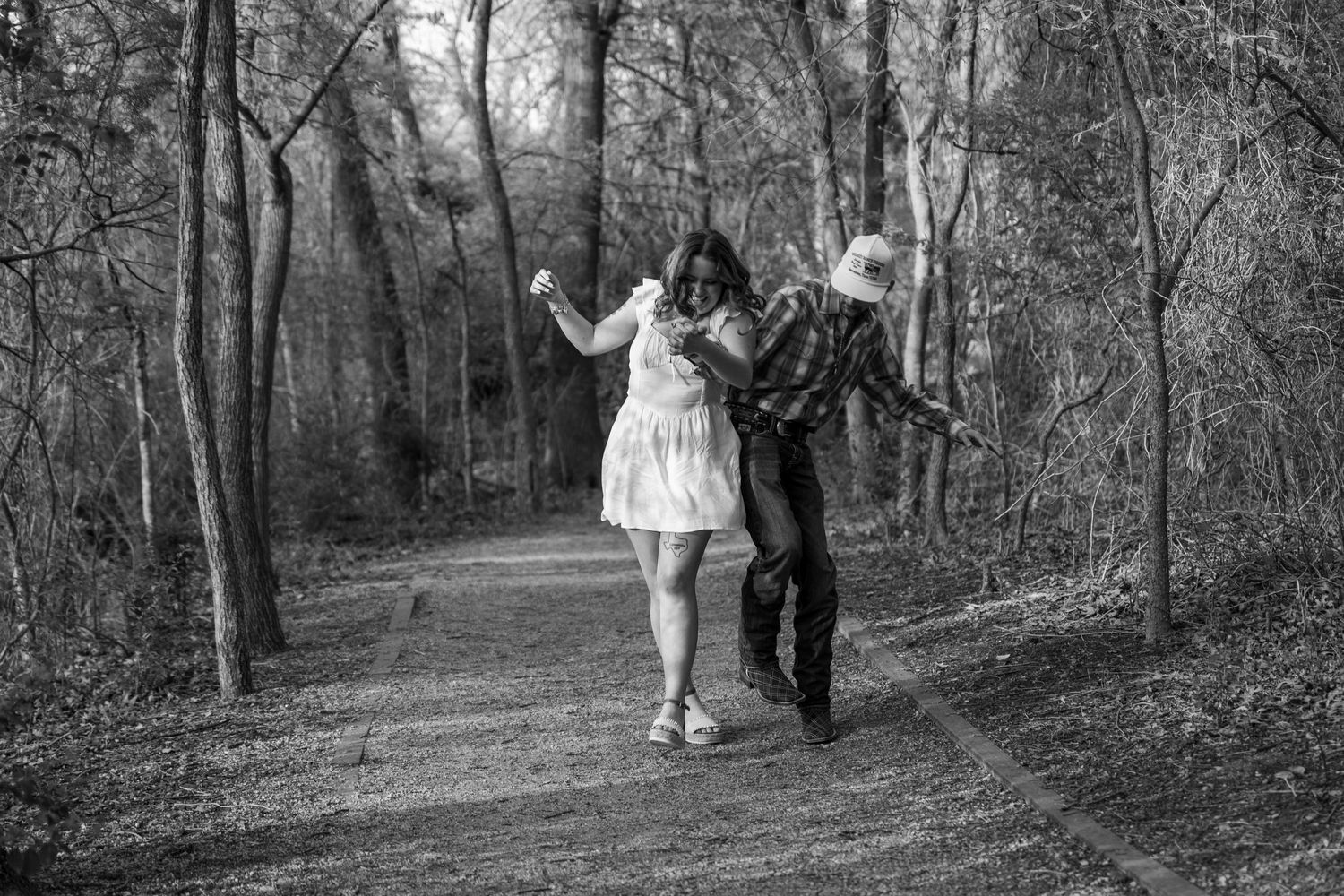 A playful black and white photo of a couple dancing on a wooded path.