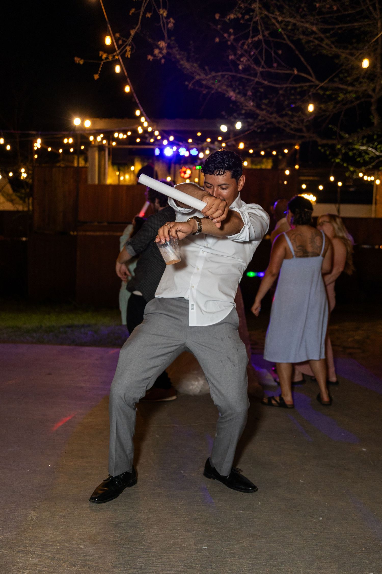 Groomsmen dancing and celebrating at outdoor evening wedding reception under string lights.