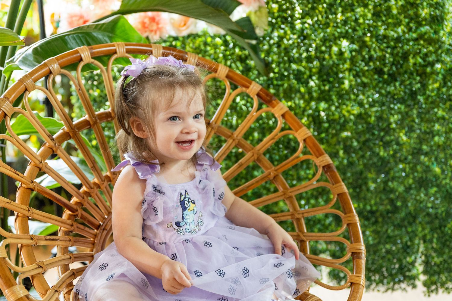 Child in floral dress sits in wicker chair against green leafy backdrop.