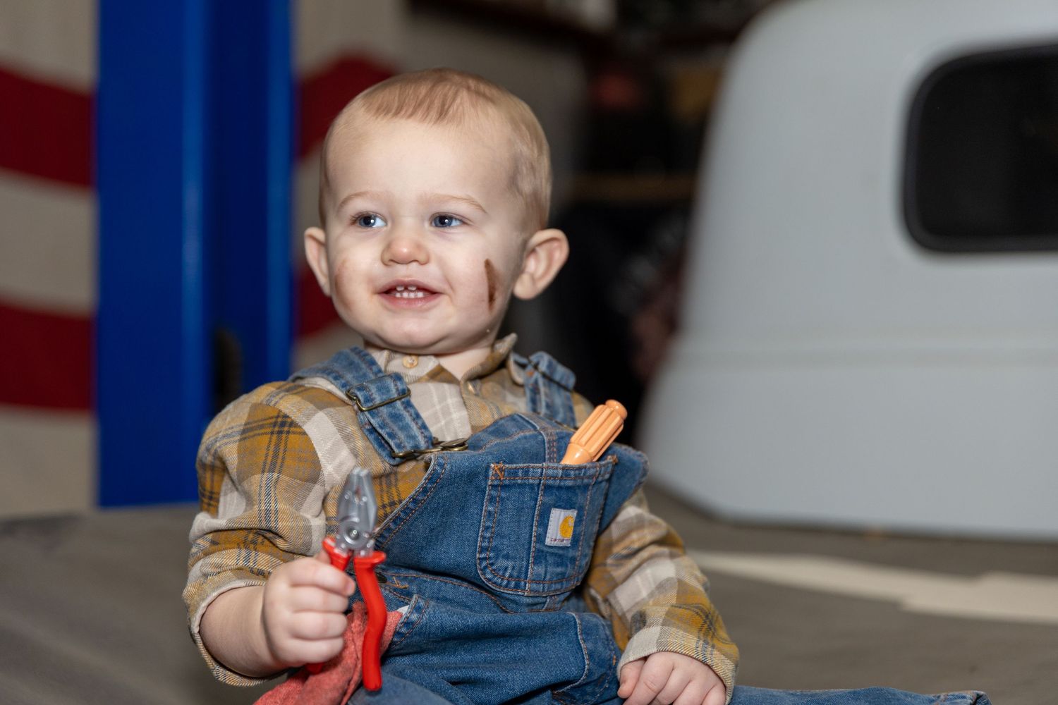 Baby in denim overalls holds tools while sitting on classic car for automotive themed photoshoot.