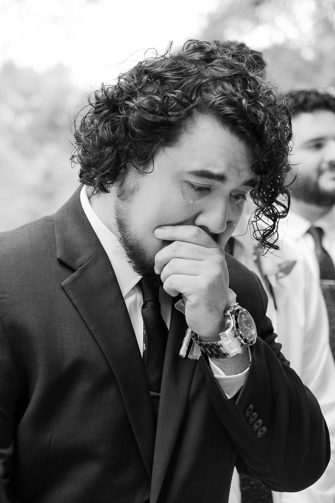Emotional groom wipes tears during wedding ceremony wearing dark suit.