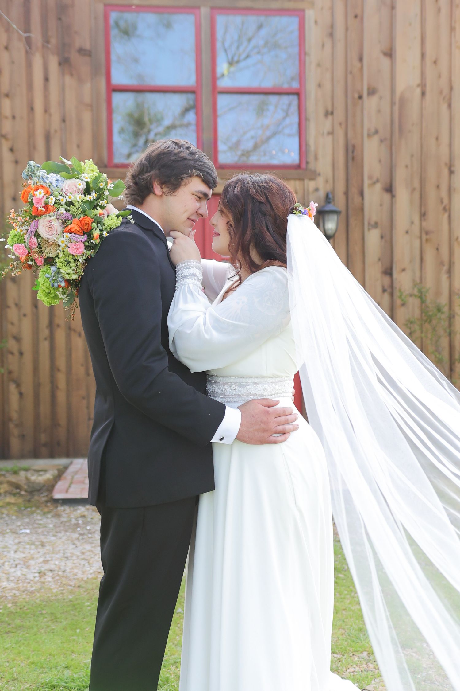 Wedding couple embracing in front of rustic wooden building with flowing white veil.
