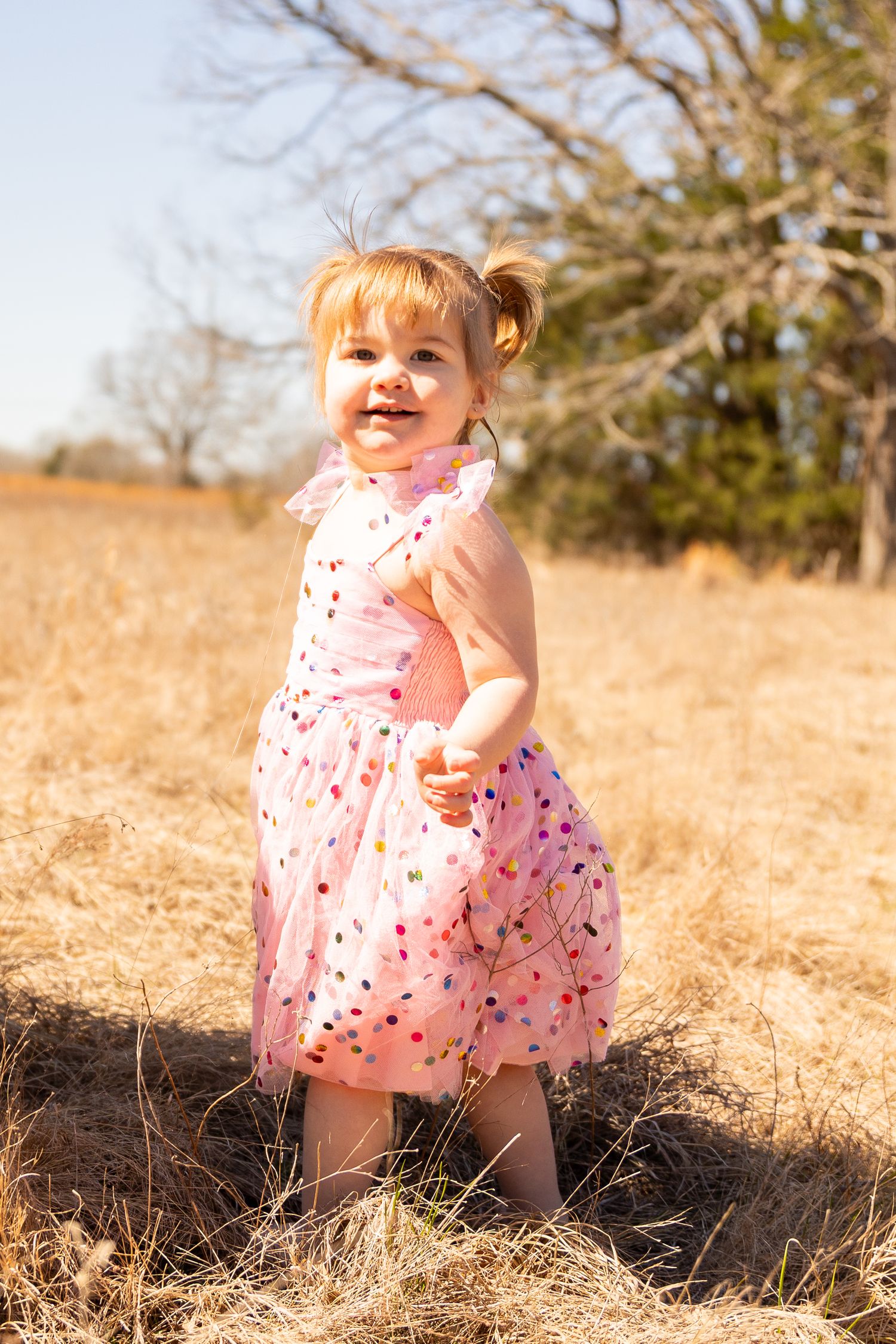 Child in pink patterned dress explores dry grass field during golden hour.