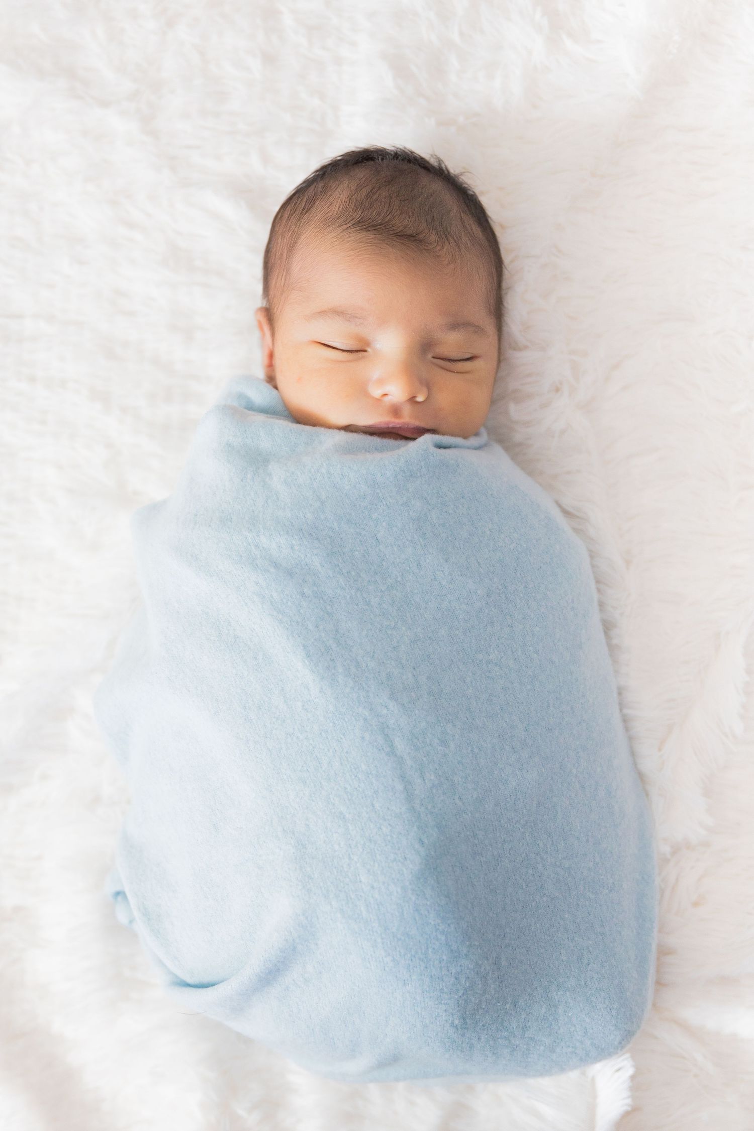 Newborn baby wrapped in soft blue blanket sleeping peacefully on white bedding.