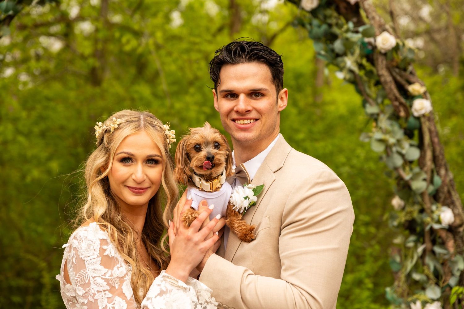 A wedding couple holds their small dog while posing under trees with floral decorations.