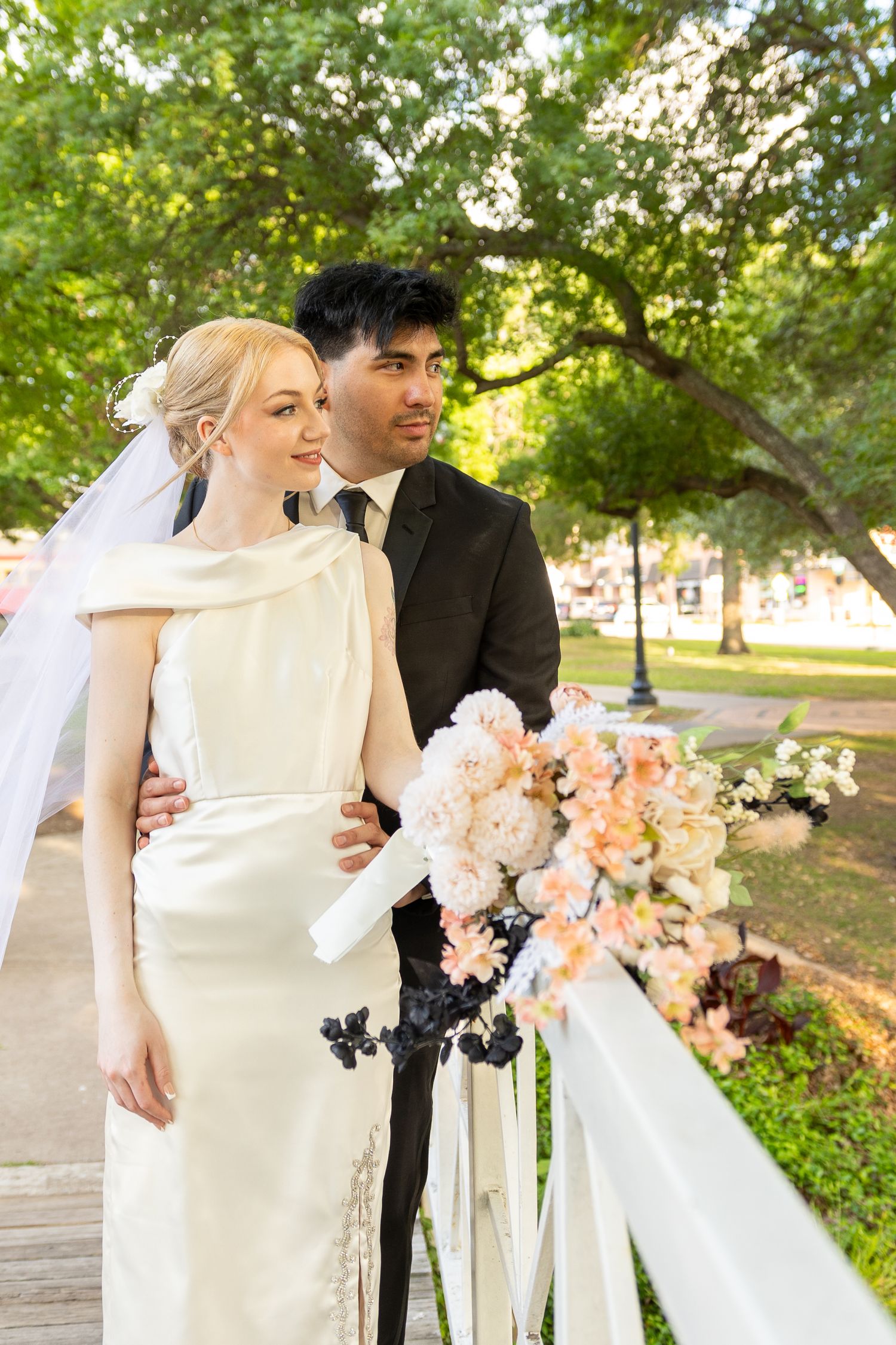 A couple walks through a garden holding peach and white wedding bouquets in soft evening light.