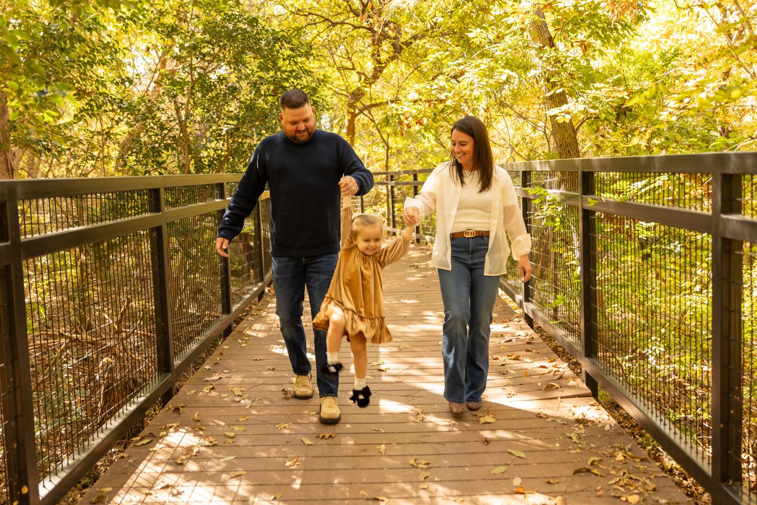 Family walking together on wooden bridge path surrounded by autumn trees.