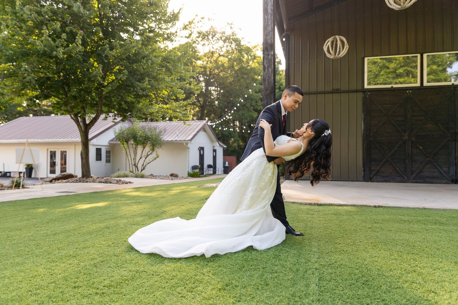 Wedding couple poses on expansive lawn with rustic barn in background at sunset.