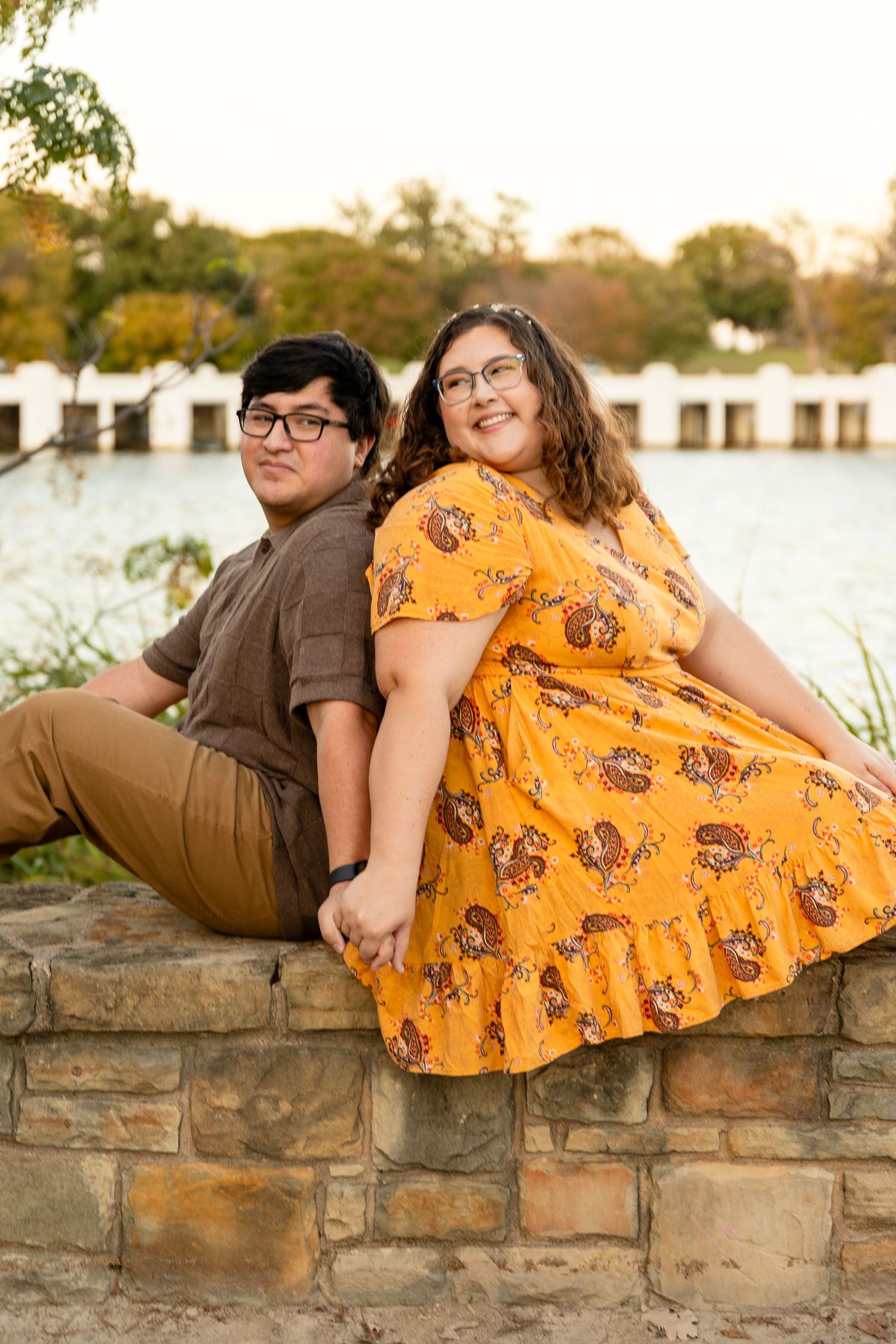 Couple relaxing on stone wall by waterfront during golden hour photoshoot.