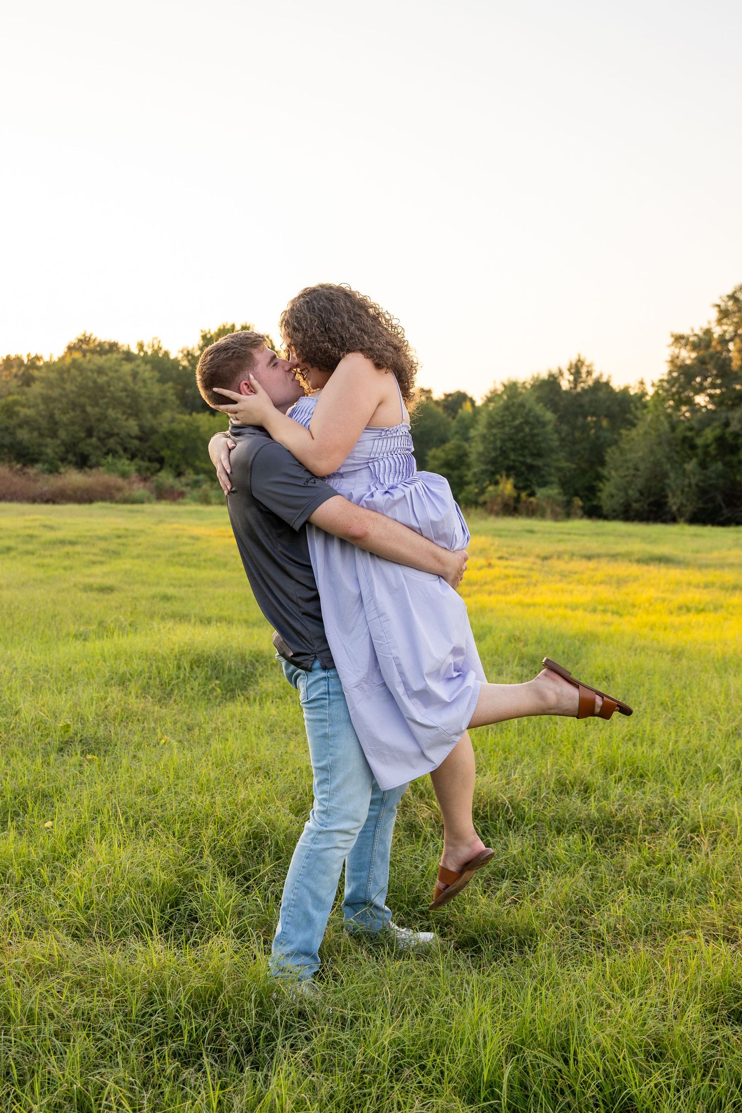 A playful lift between a couple in a sundress and casual wear in a grassy field.