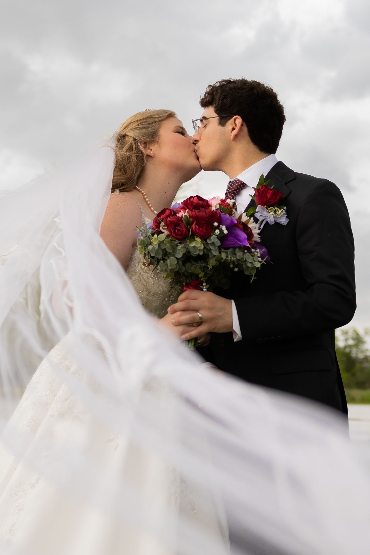 Romantic wedding couple embracing outdoors with flowing veil and dramatic cloudy sky.