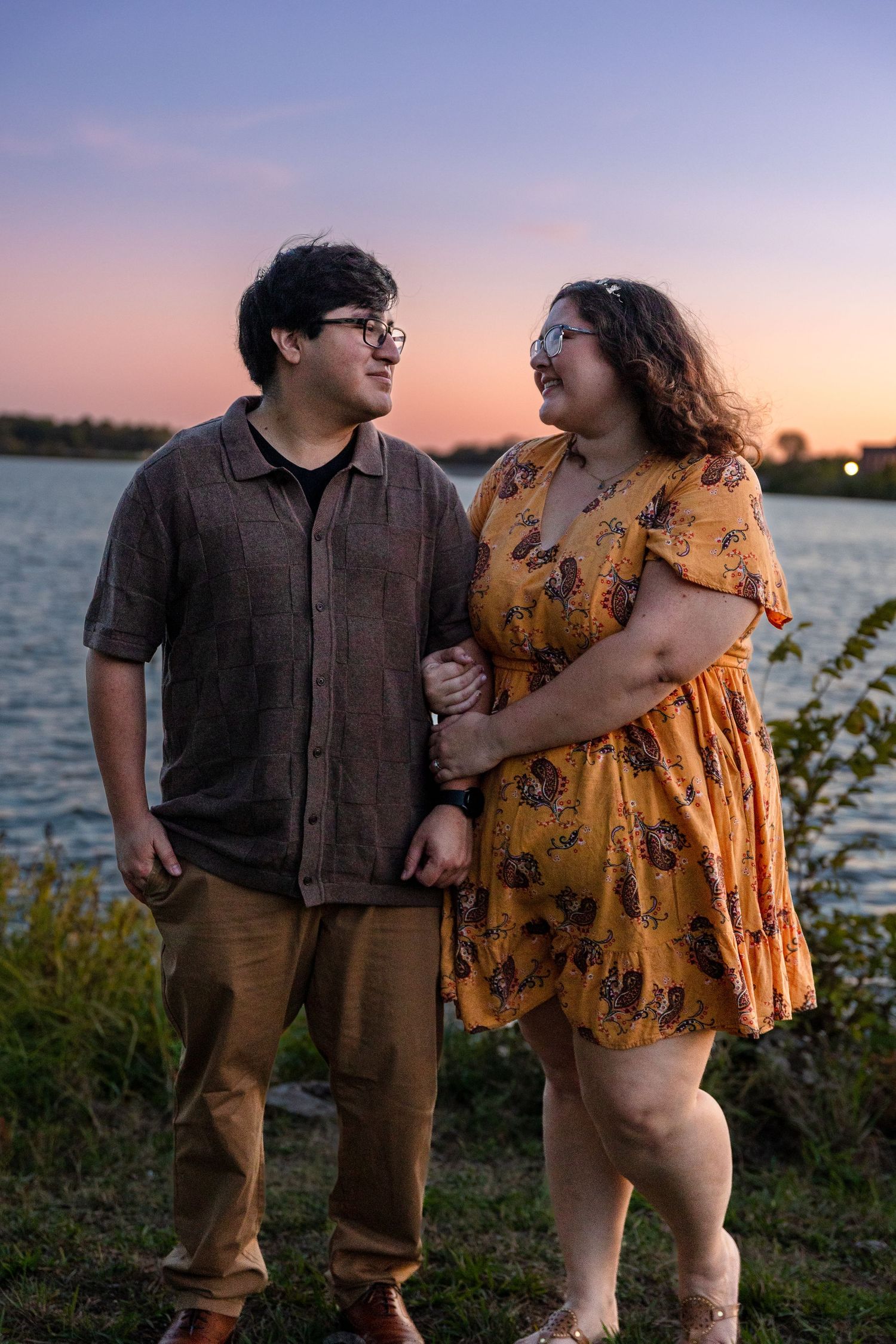 Romantic couple in casual attire posing by the waterfront during sunset.