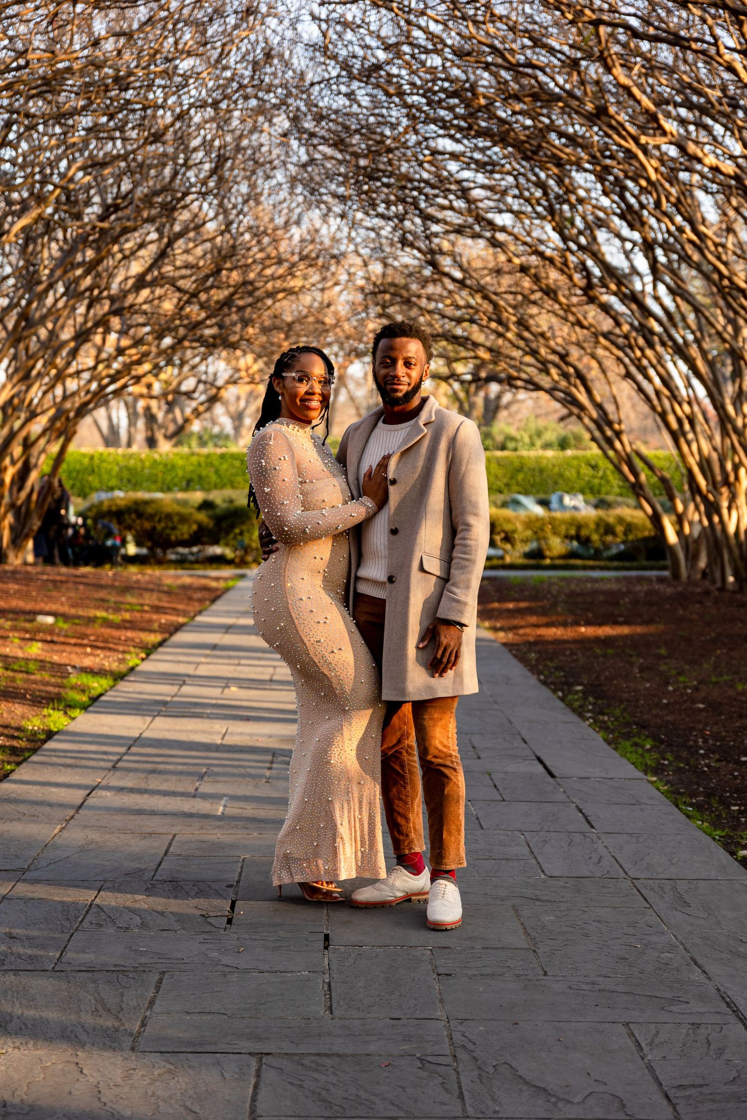 A stylish couple poses under an archway of bare branches during golden hour.