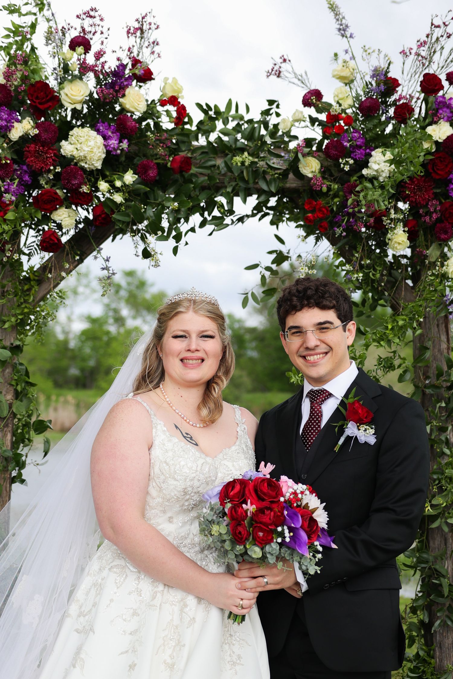 Wedding ceremony under a floral arch with deep red and purple blooms against a lush outdoor backdrop.