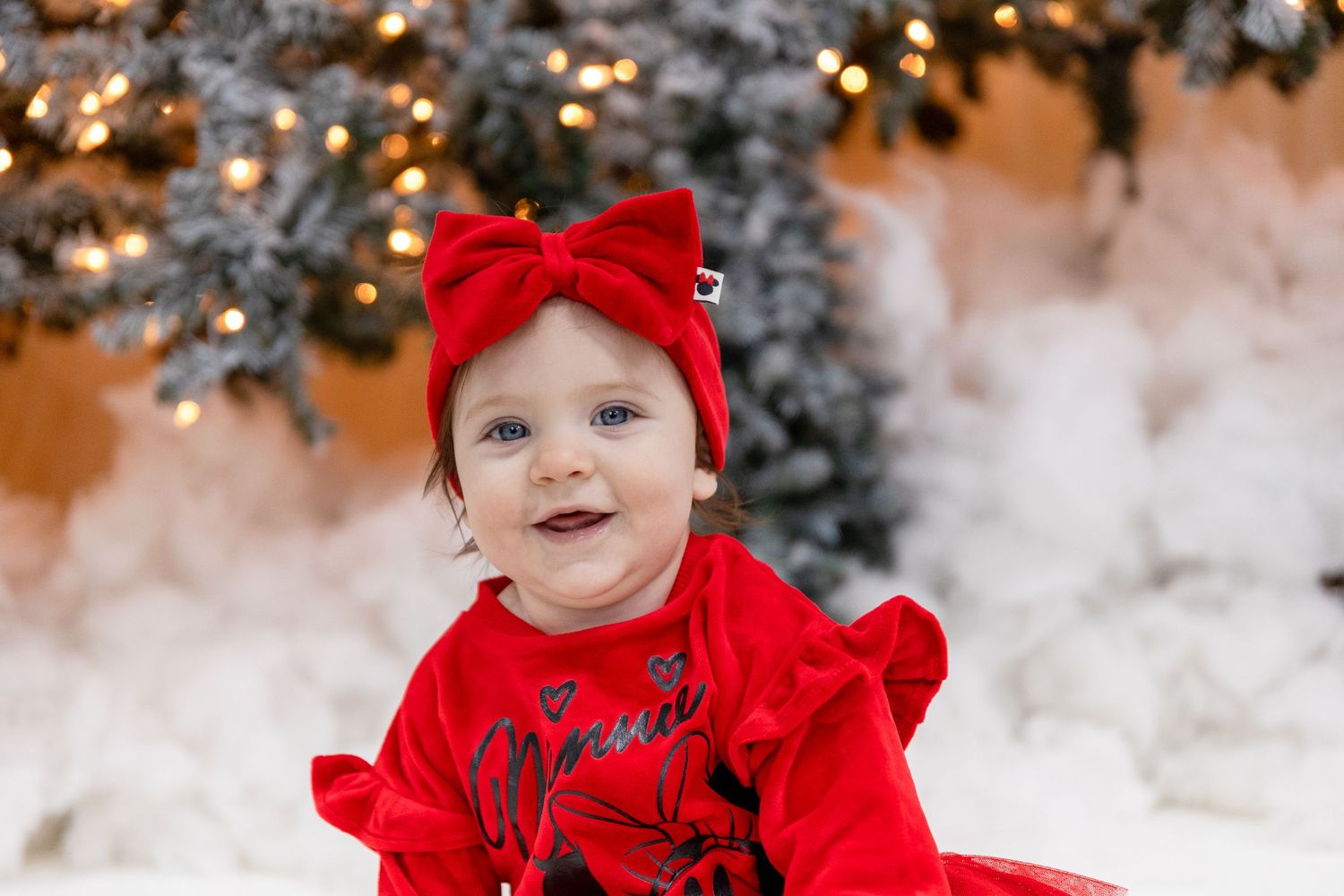 Baby in bright red holiday outfit poses among snowy Christmas decorations.