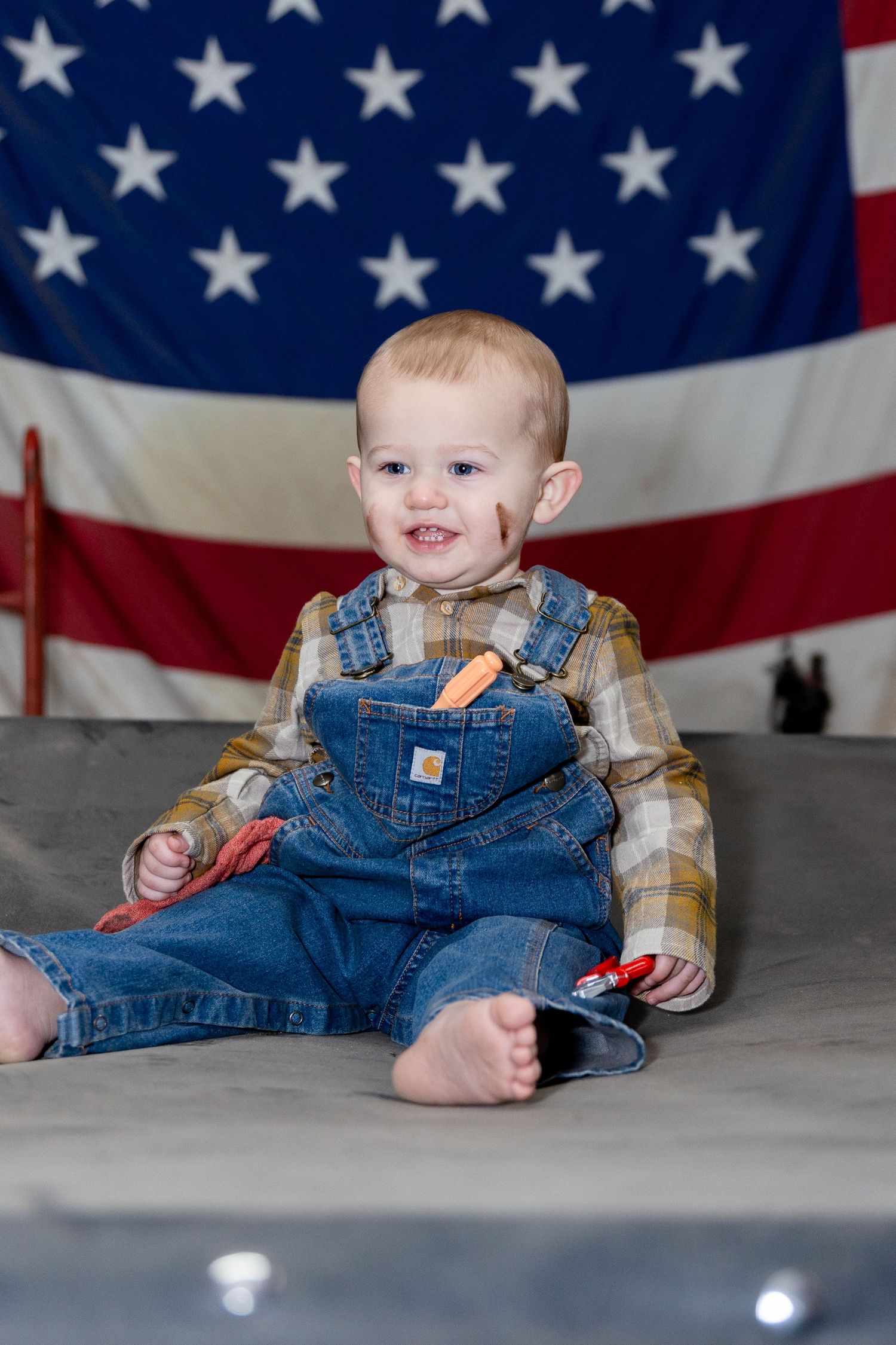 Baby wearing denim overalls poses against American flag backdrop for patriotic photo session.