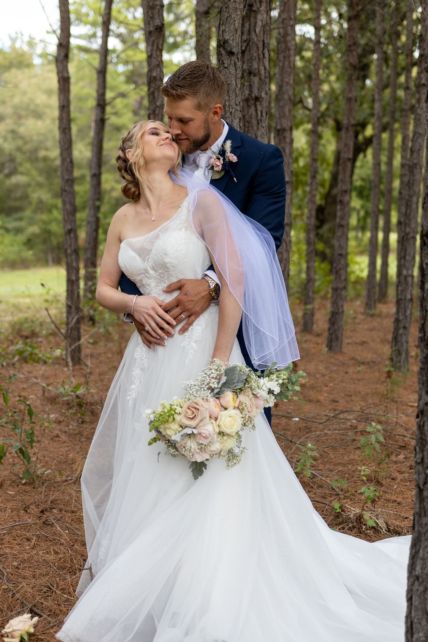 Wedding couple embracing among pine trees with white floral bouquet.