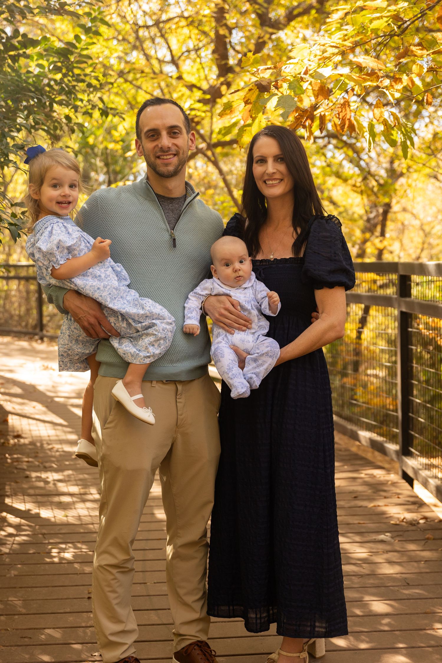 Family portrait session on wooden bridge with golden autumn trees in background.