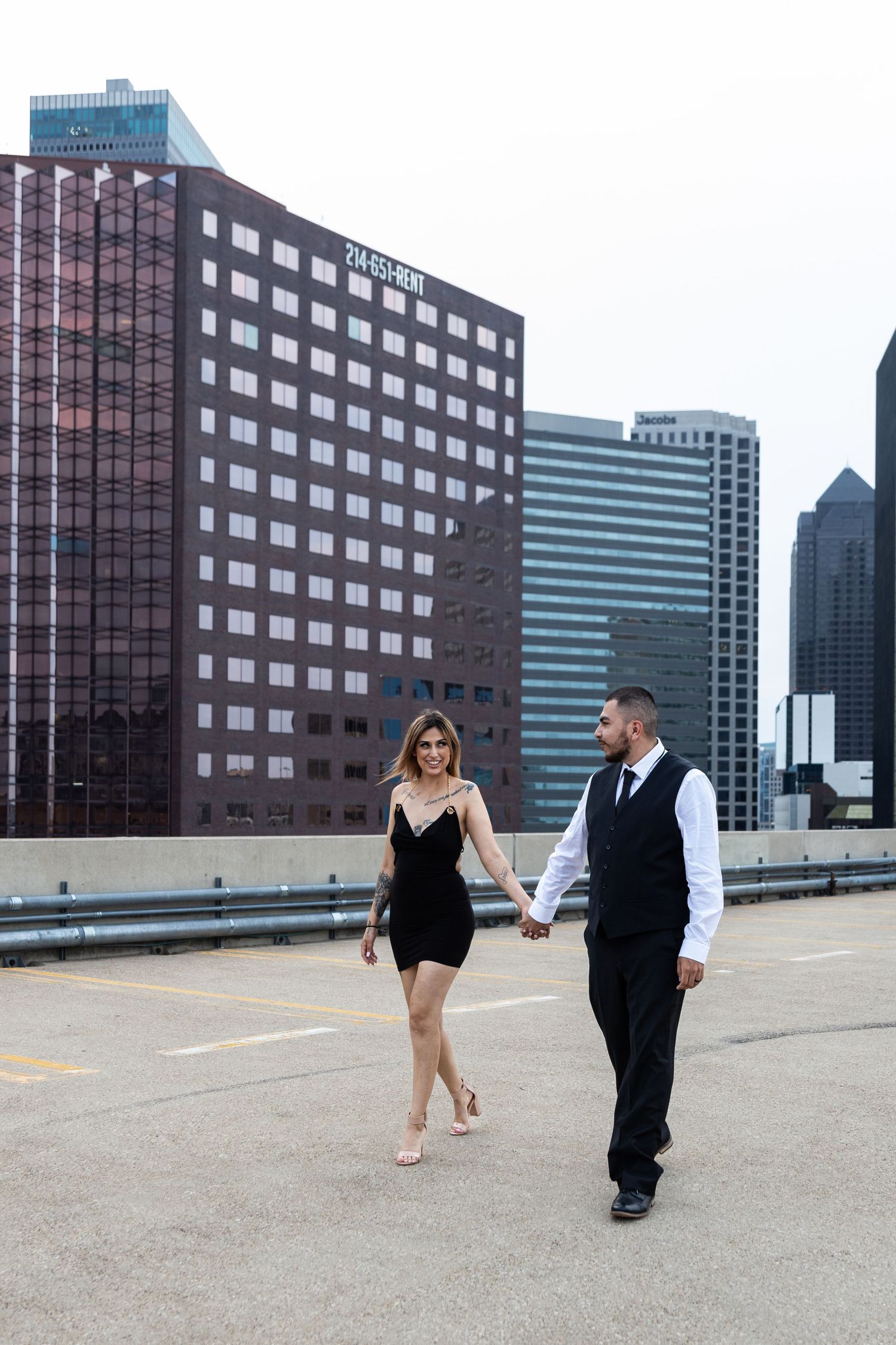 Couple in formal wear walks playfully across urban rooftop with city skyline backdrop.