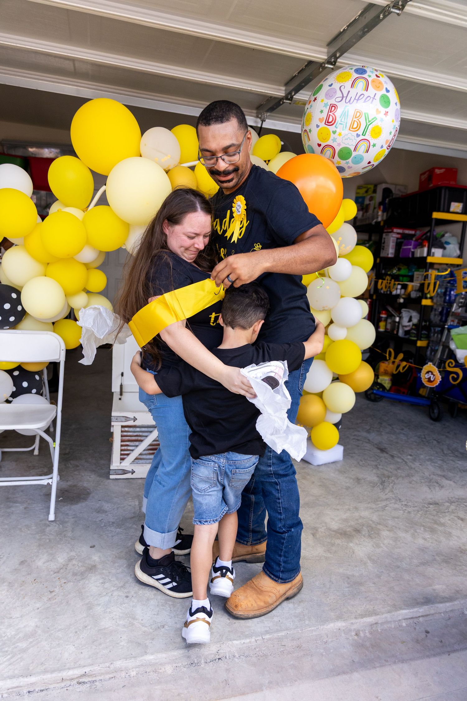 Family celebrating with yellow and orange balloon arch decorations in garage setting.