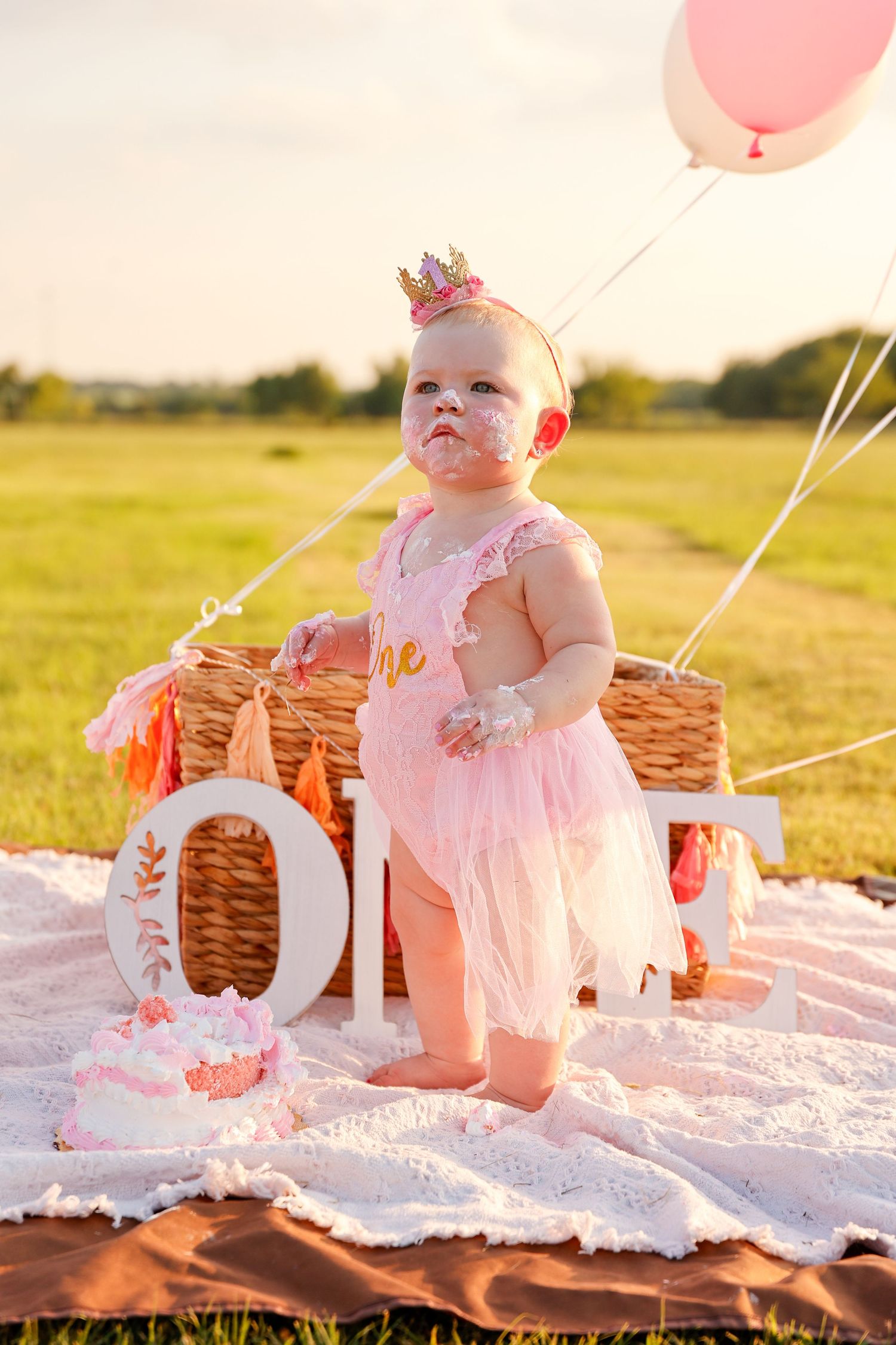 Baby in pink dress sits on white blanket with pink balloons and wicker basket during golden hour photoshoot.