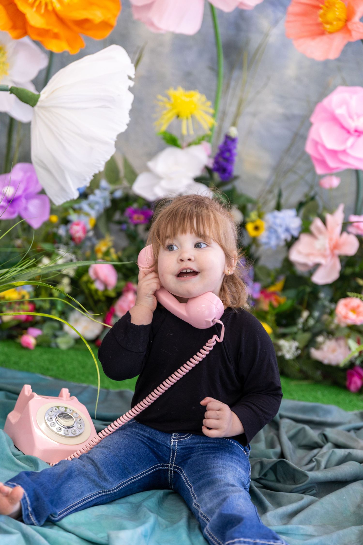 Child in black sweater plays with pink retro telephone among colorful spring flowers.
