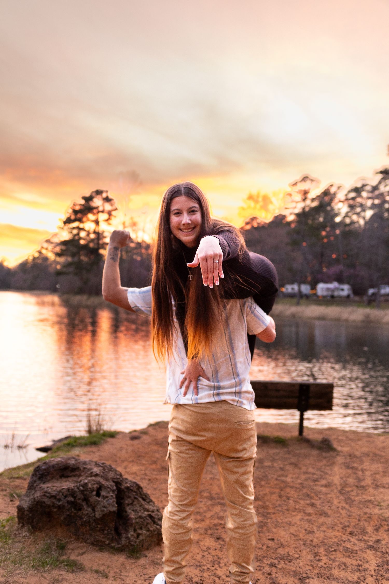 A couple takes engagement photos by a lake during golden hour with trees in the background.