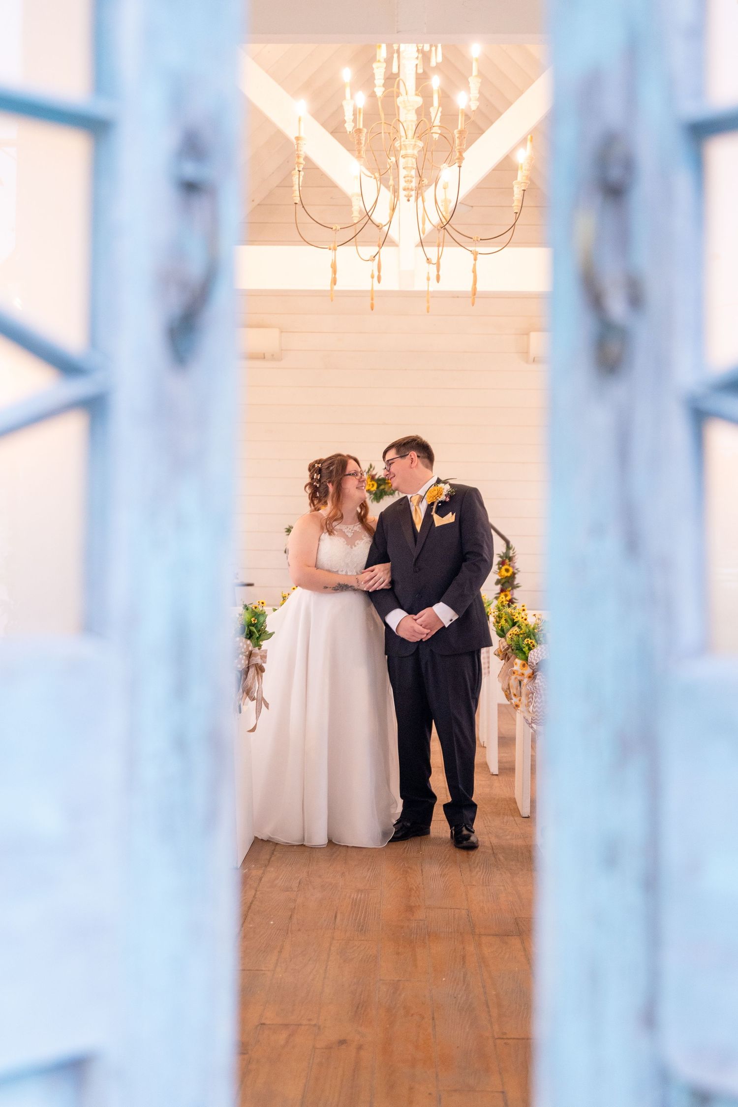 A wedding couple shares a romantic moment in an elegant white room with hardwood floors and chandeliers.