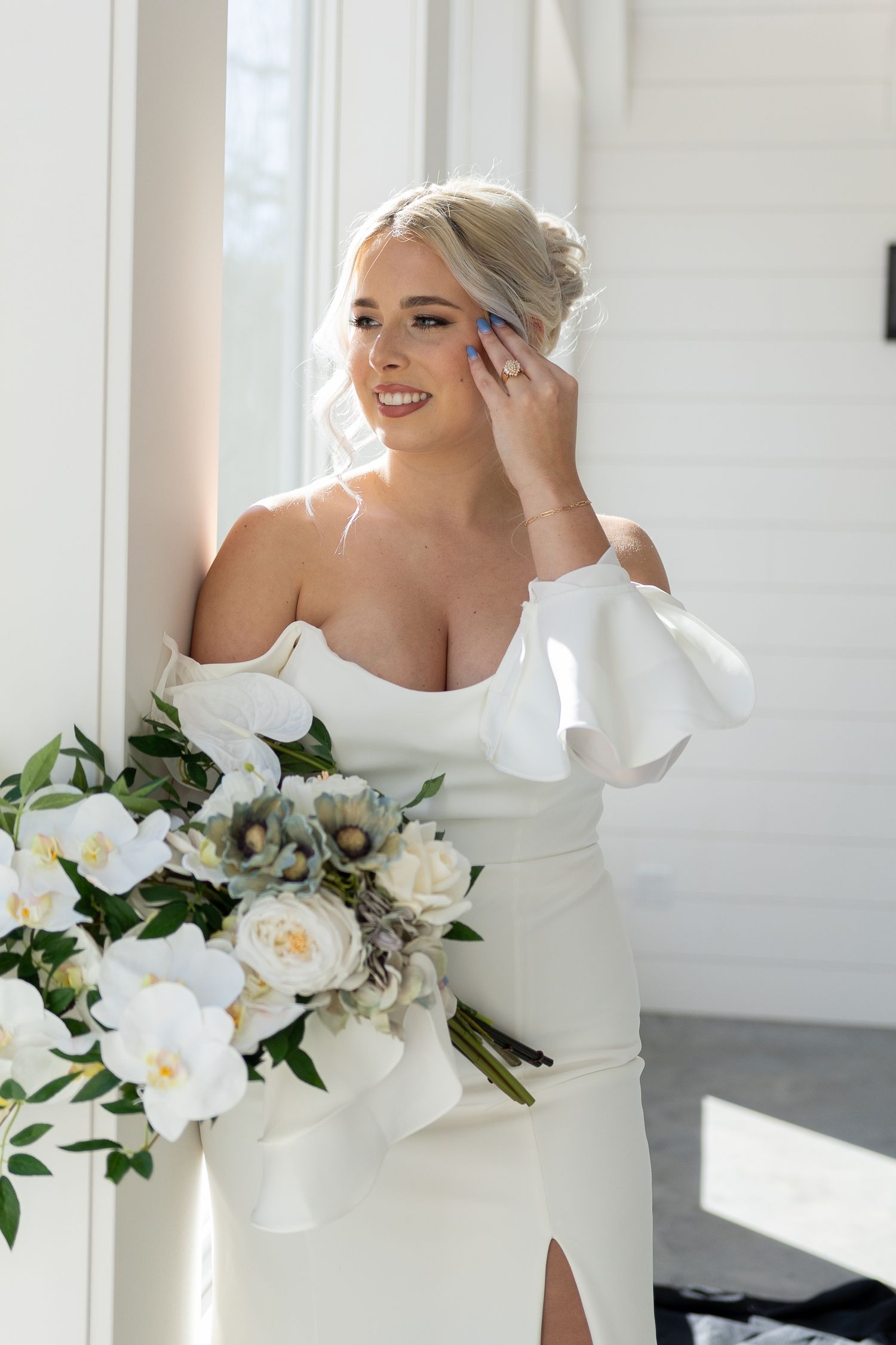 Bride in elegant off-shoulder white dress holding a bouquet of white roses and greenery in a bright sunlit room.