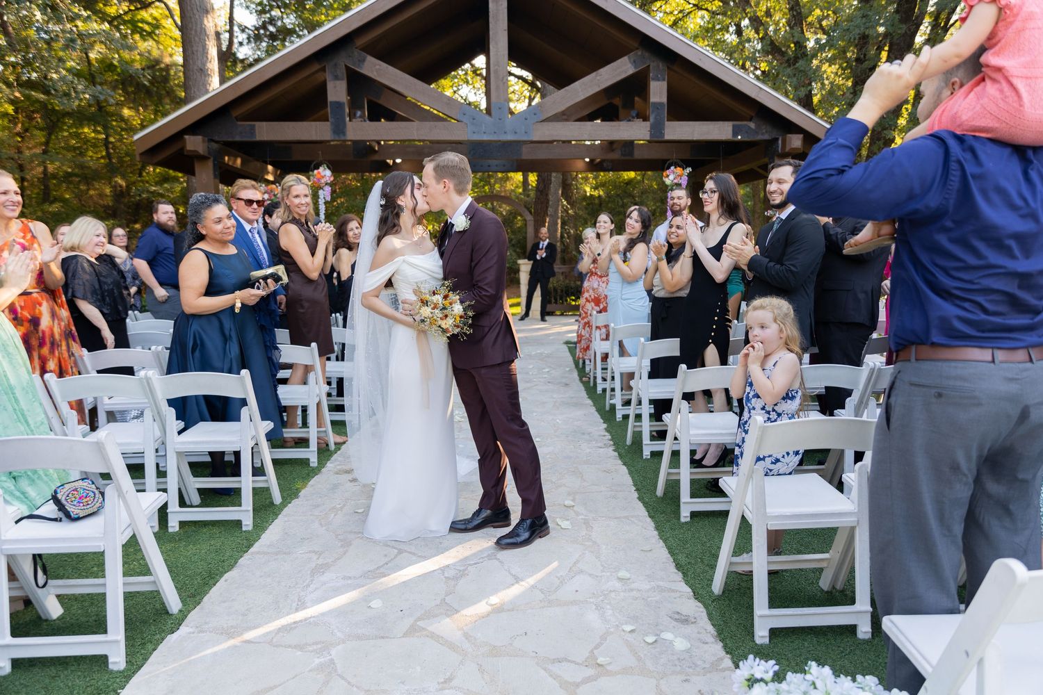 Wedding ceremony taking place under a wooden pavilion with white chairs and guests in attendance.