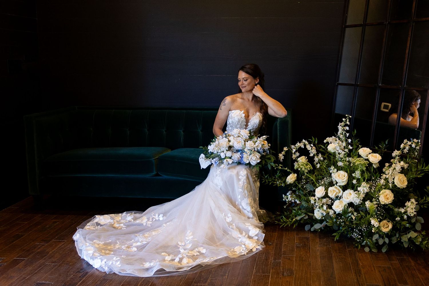 A bride in an elegant lace gown poses dramatically with floral arrangements in a dark venue.