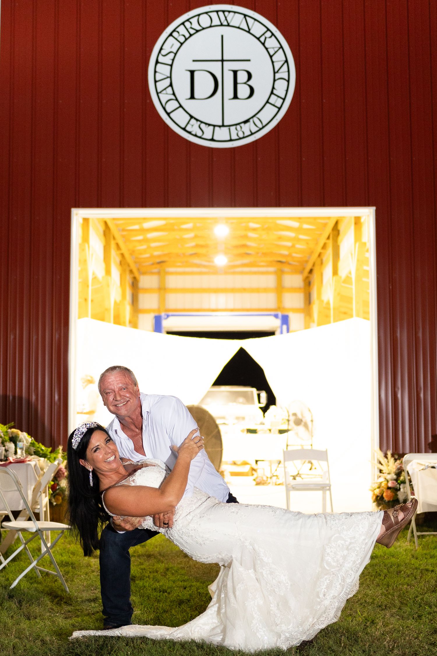 Wedding couple poses playfully outside red barn with illuminated DB logo above.