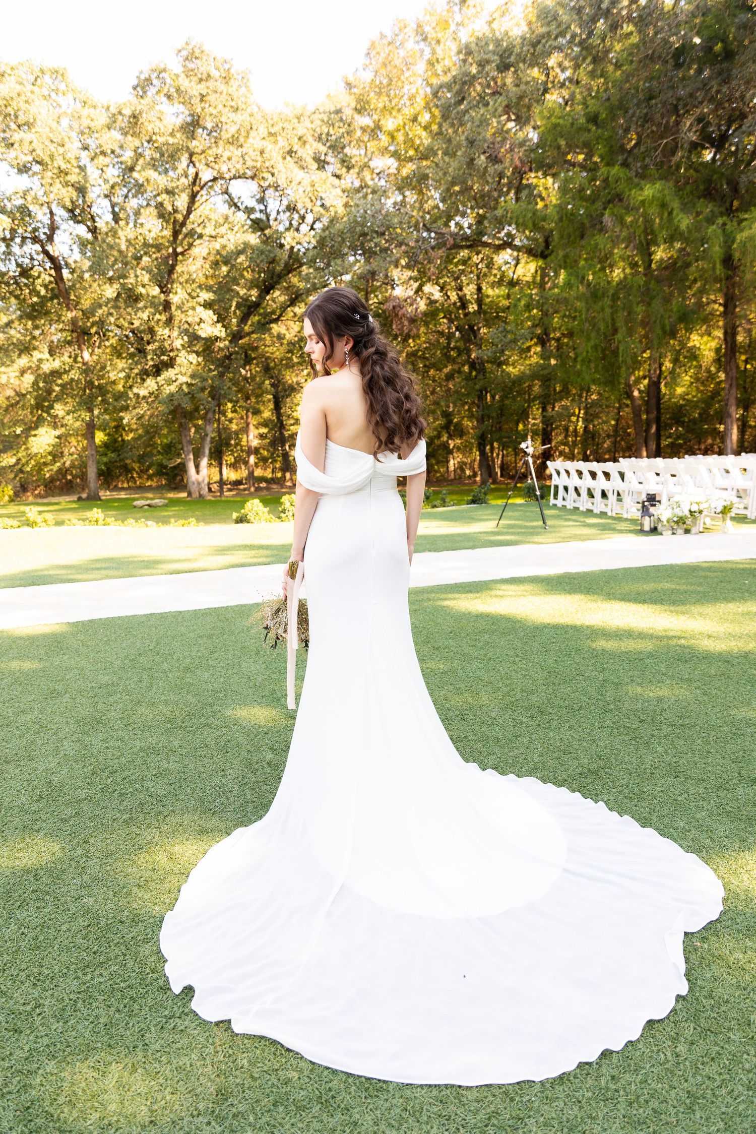 A bride in a flowing white wedding dress with long train stands on a manicured lawn surrounded by trees.