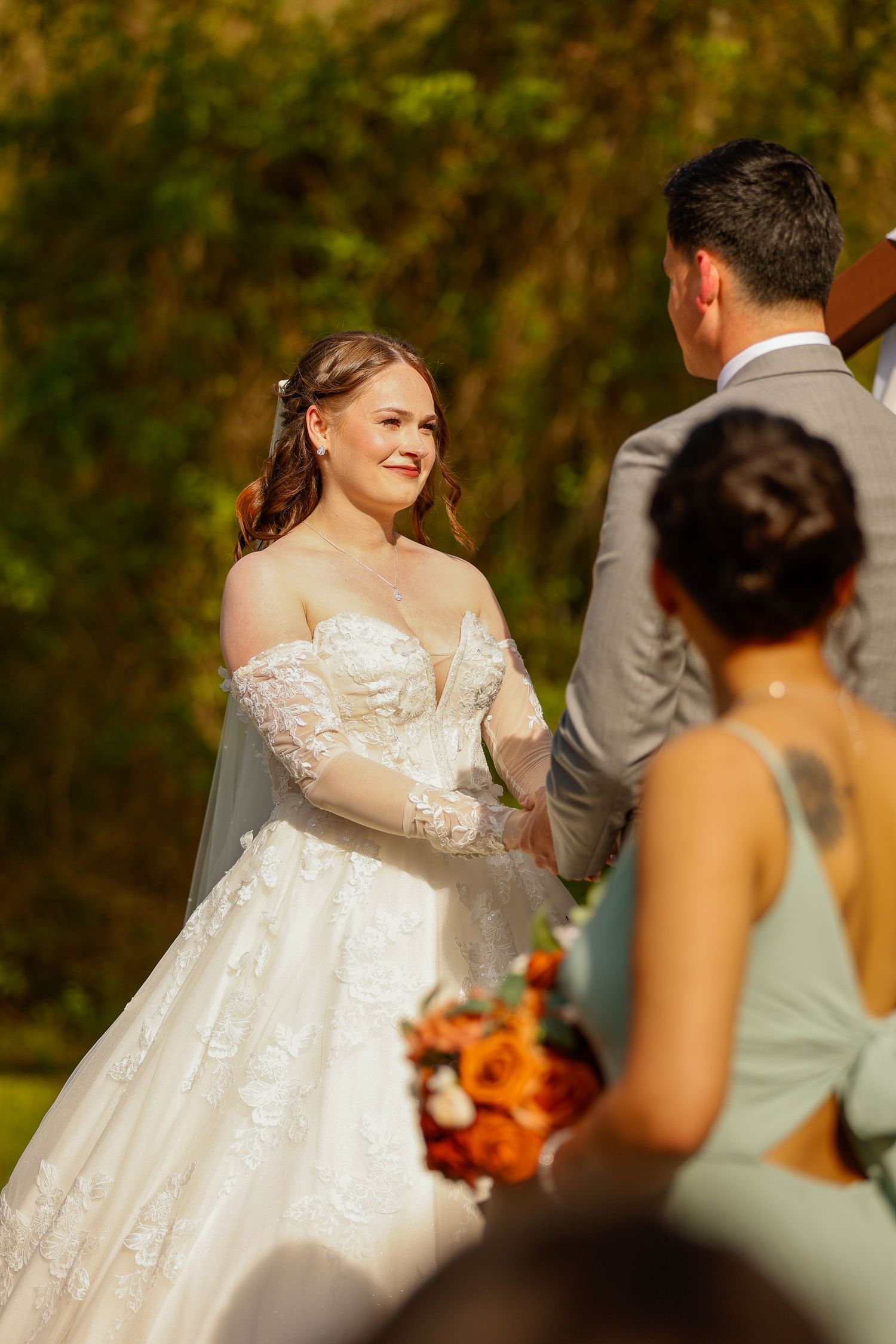 A bride smiles during her outdoor wedding ceremony with fall foliage in the background.