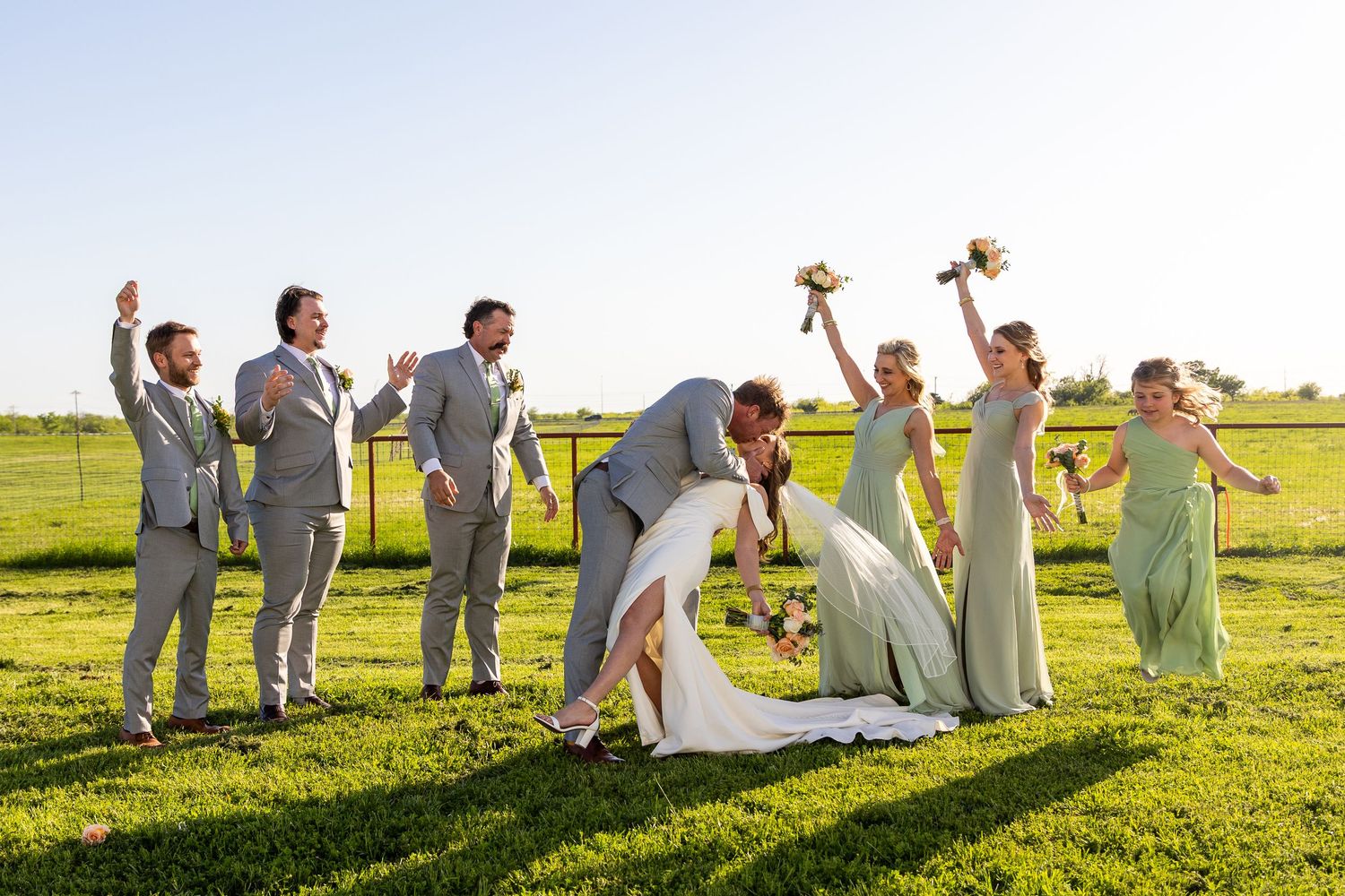 Wedding party poses joyfully on green lawn during sunset celebration.