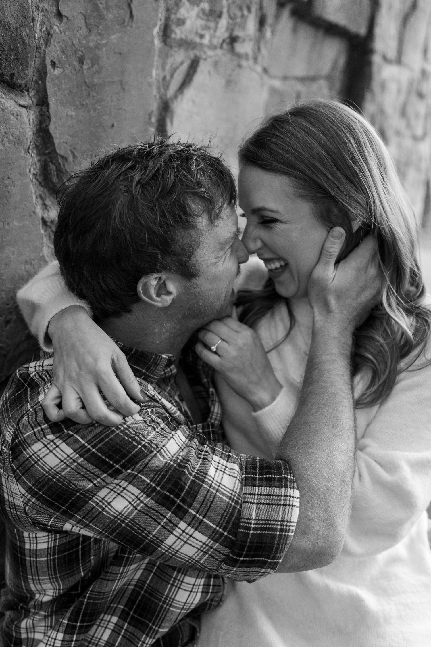 A black and white candid photo capturing a joyful moment between a couple against a stone wall.