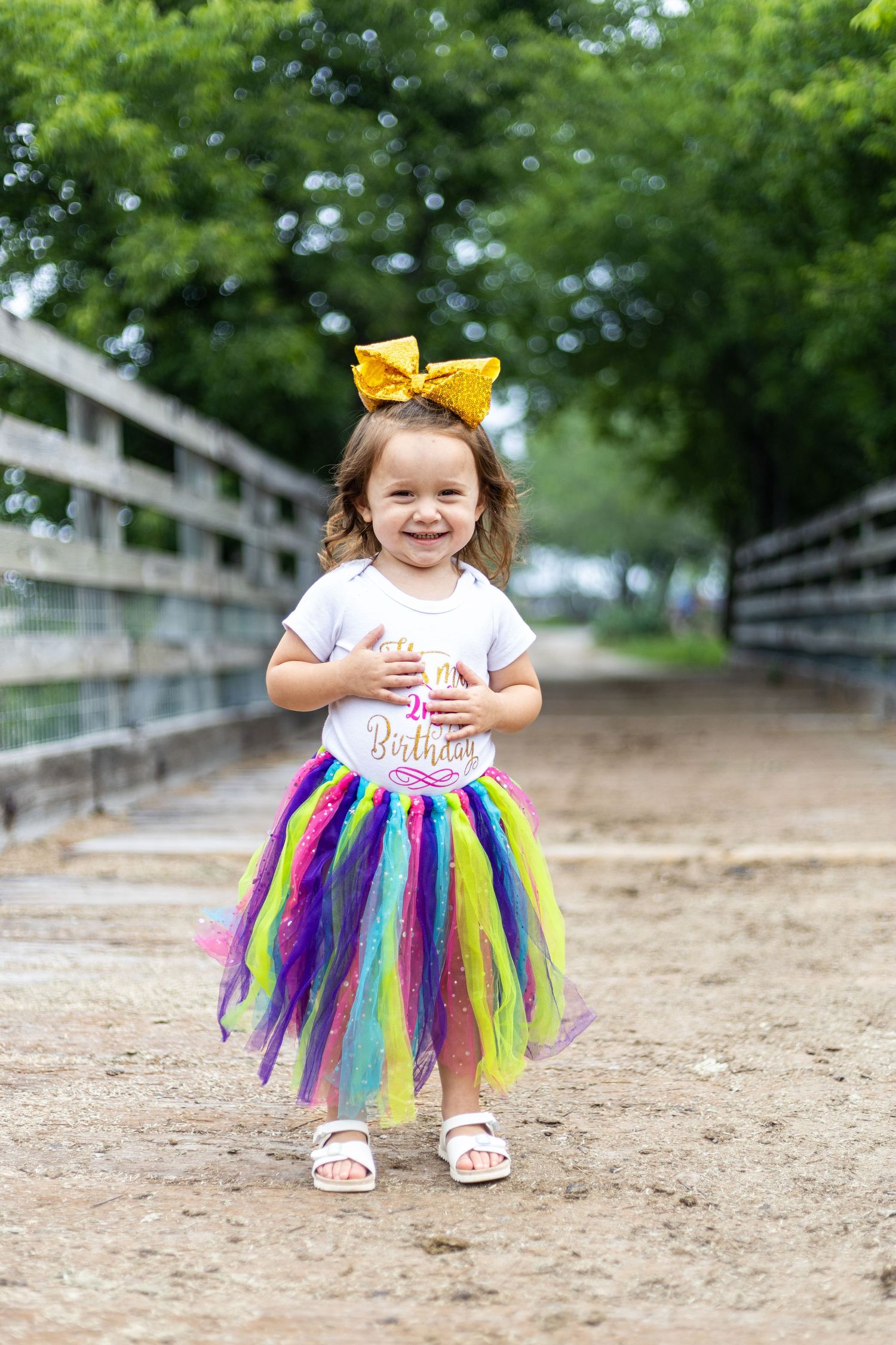 Child wearing rainbow tutu and white shirt playing near wooden fence.