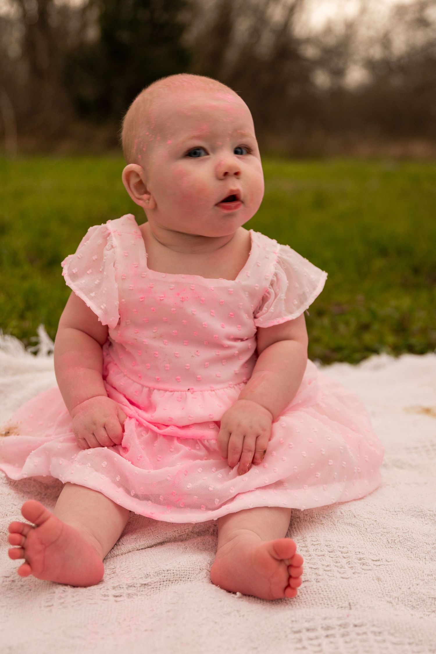 Baby in pink ruffled dress sits on white lace blanket outdoors.