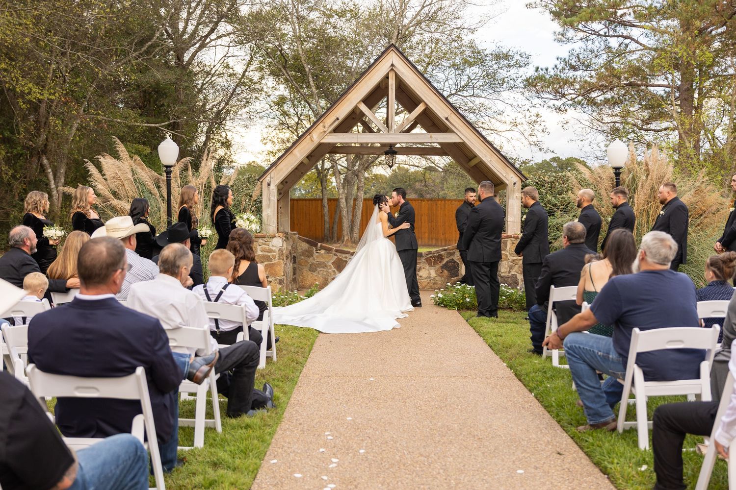 Rustic outdoor wedding ceremony with wooden arch and seated guests under tree canopy.