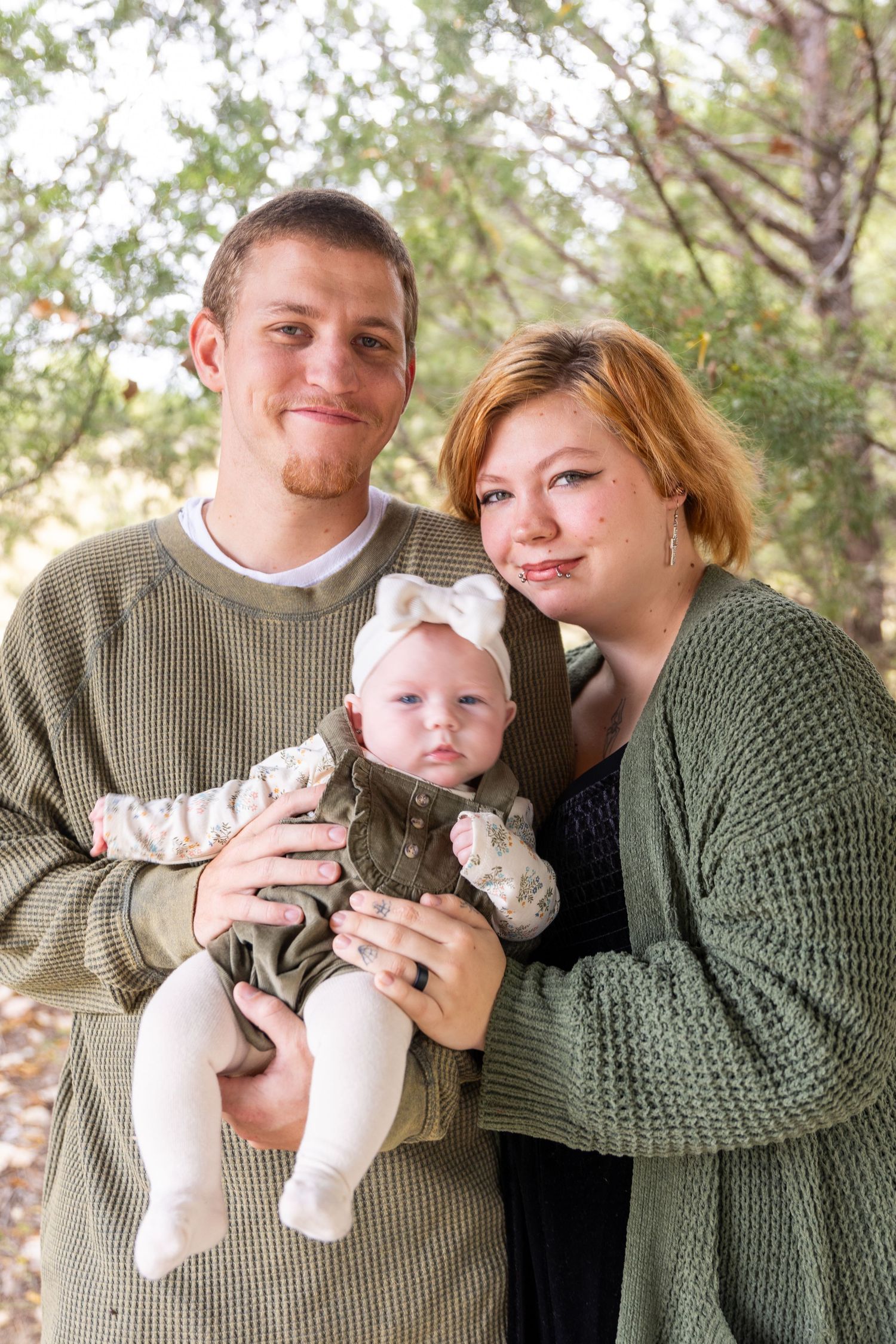 Parents in green sweaters holding and cuddling with baby in outdoor setting.