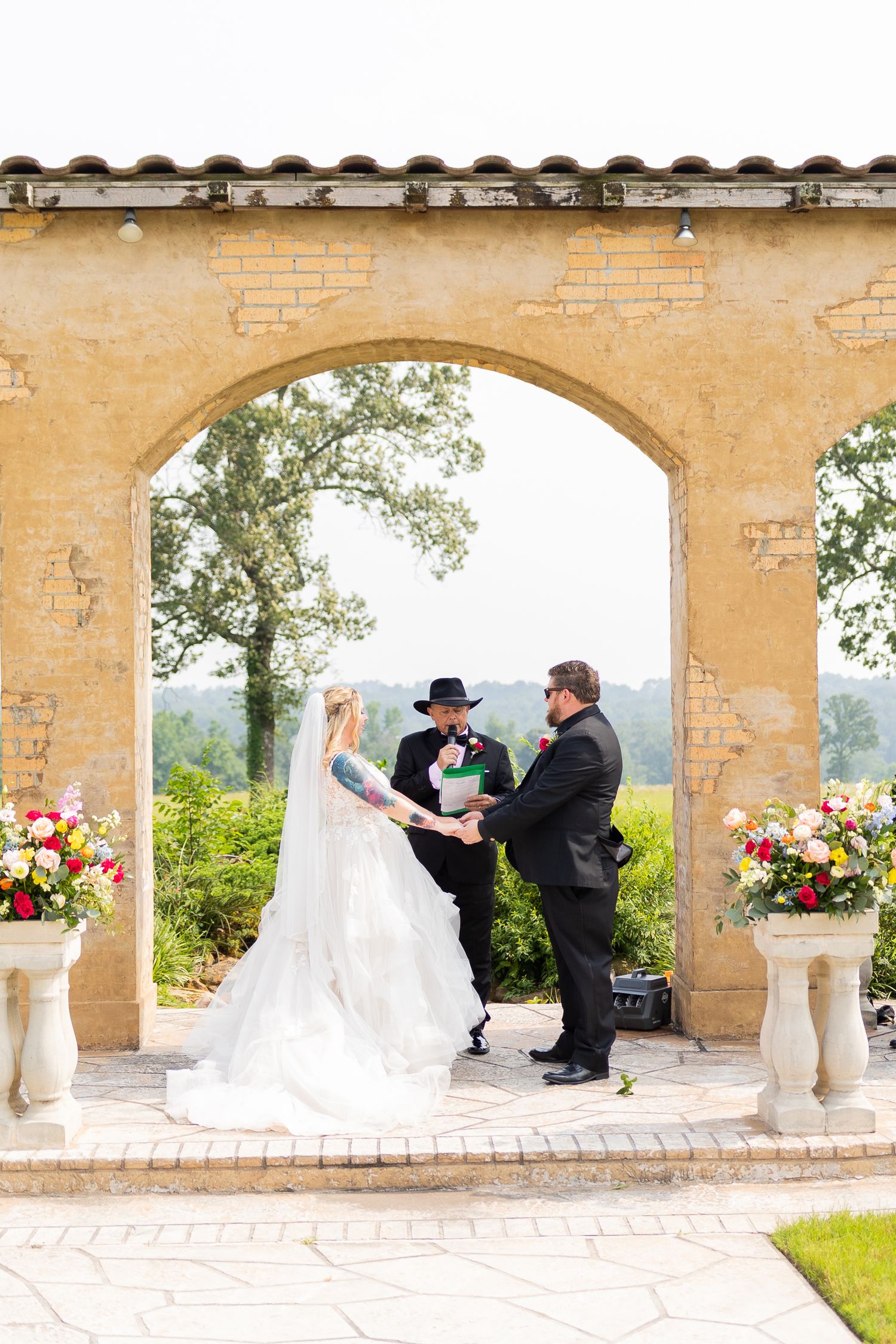 A wedding ceremony unfolds under stone arches decorated with pink floral arrangements.