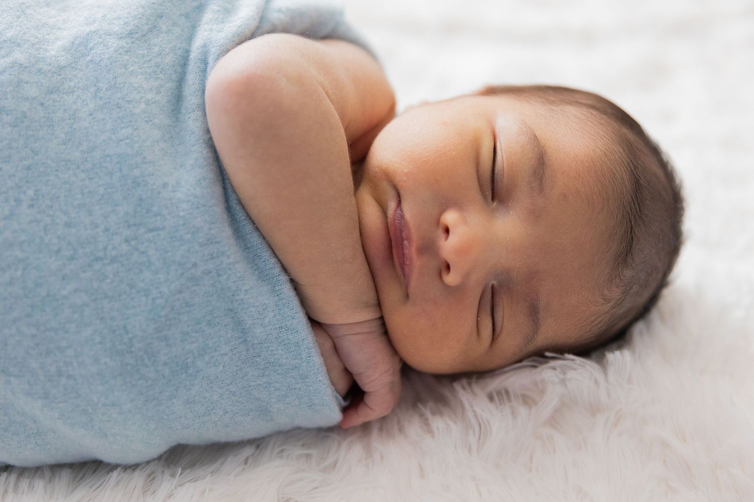 Peaceful newborn baby sleeping while wrapped in light blue swaddle on white surface.