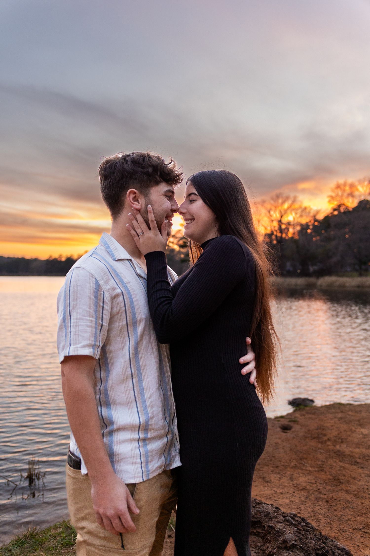 A couple embraces by a lake during a golden sunset with trees in the background.