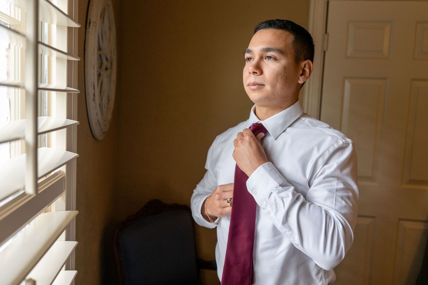Groom in white shirt and burgundy tie stands thoughtfully by window in series of candid getting ready shots.