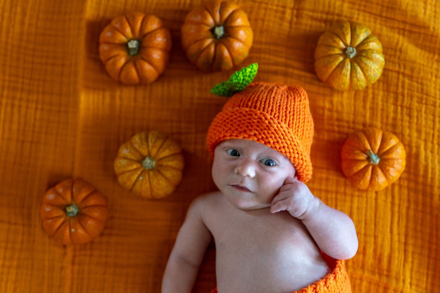 A baby wearing an orange knit pumpkin hat lying on orange fabric surrounded by mini pumpkins.
