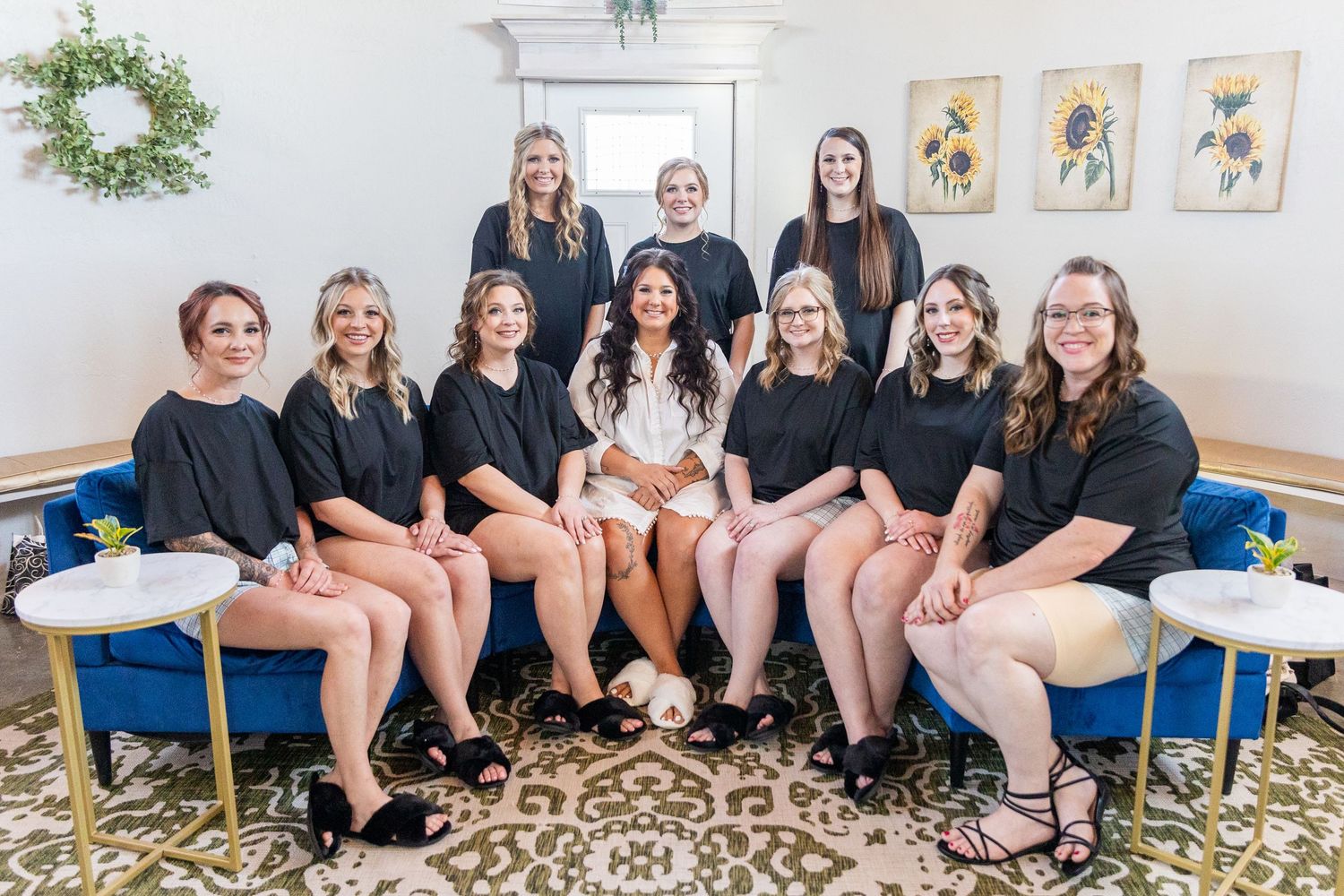 Wedding party poses on elegant cream furniture in a formal setting with botanical artwork on the wall.