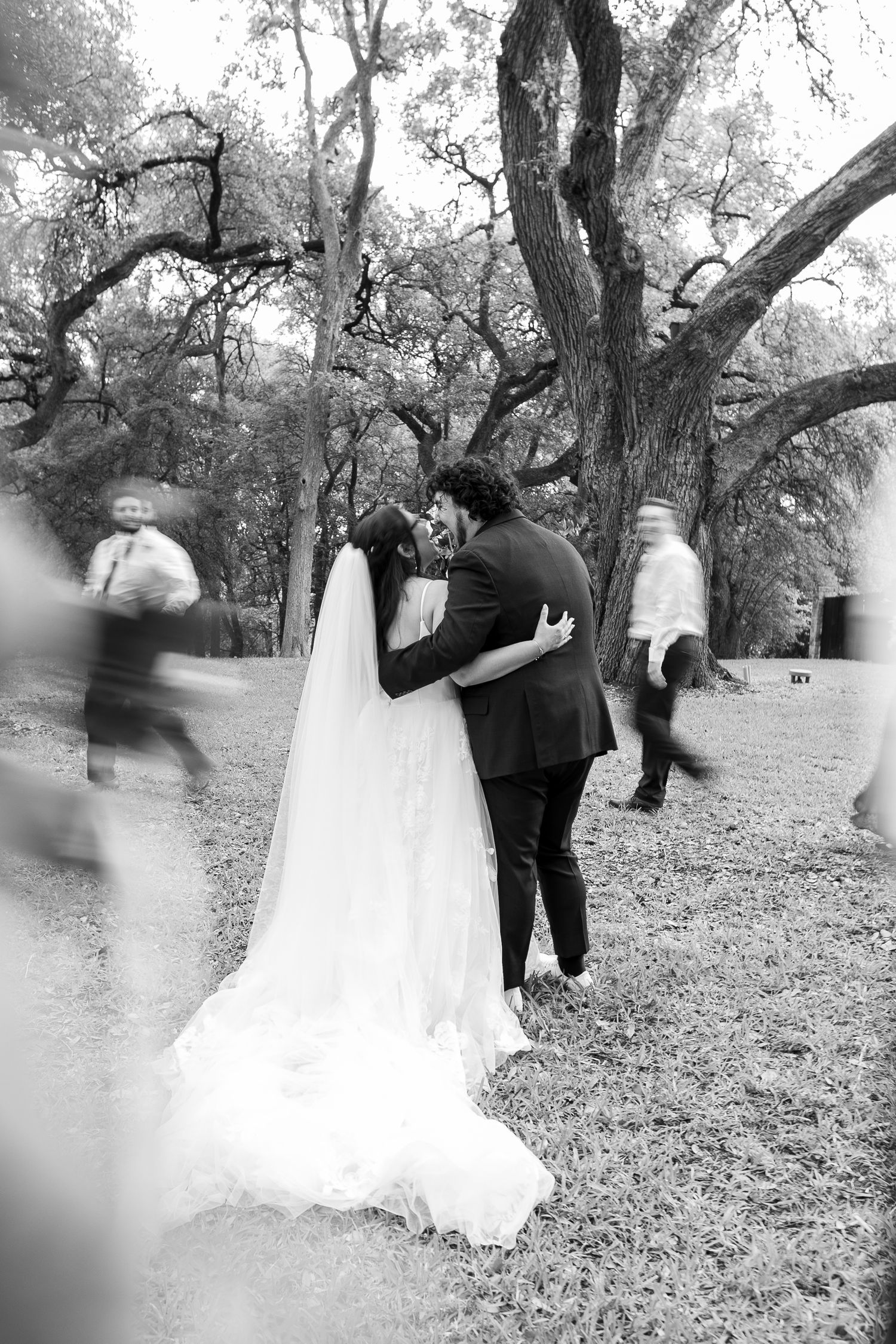 A tender moment between newlyweds sharing their first kiss under majestic oak trees in black and white.